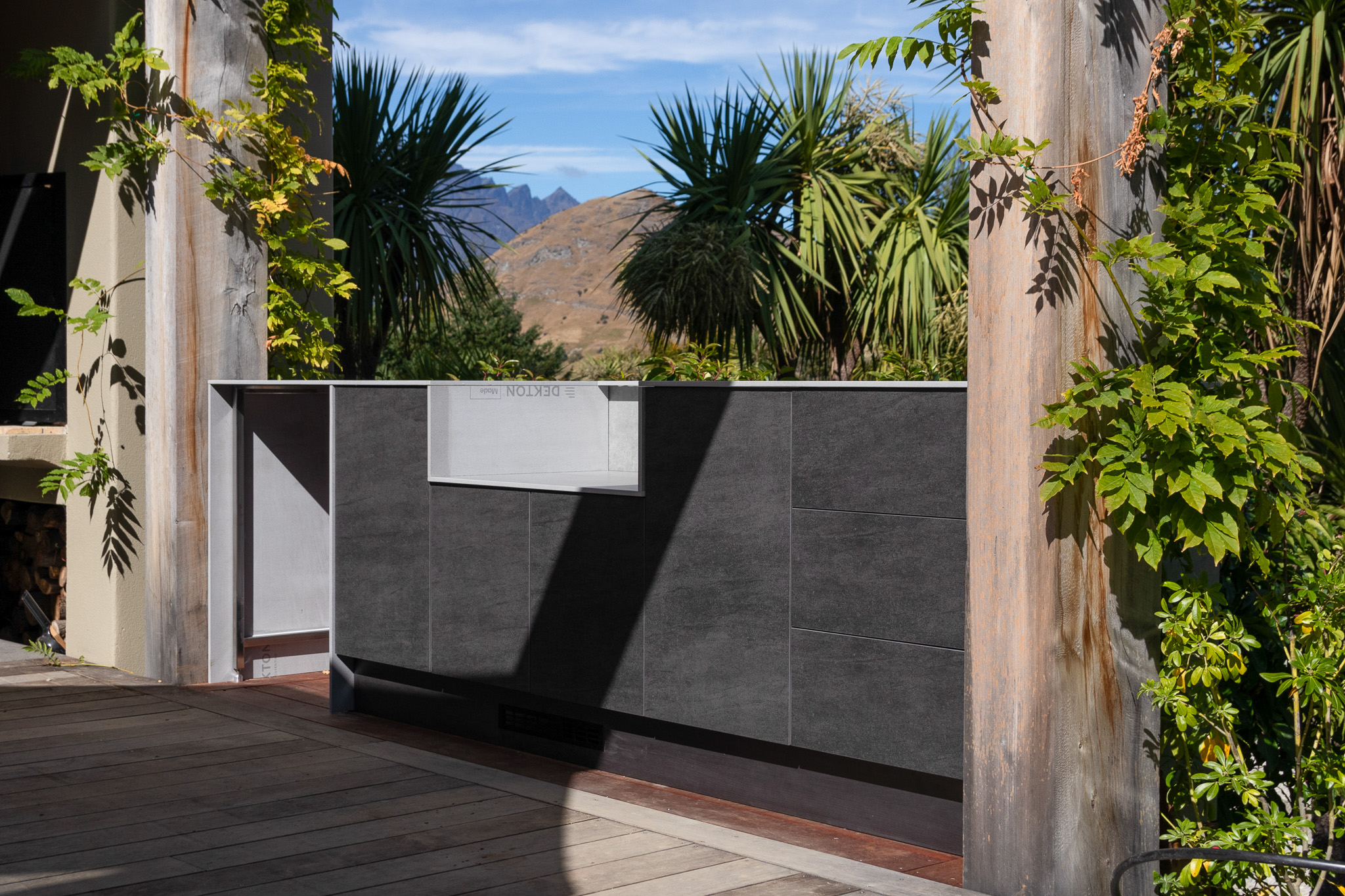 Outdoor kitchen island with dark cabinetry on a wooden deck surrounded by green plants and mountains in the background.