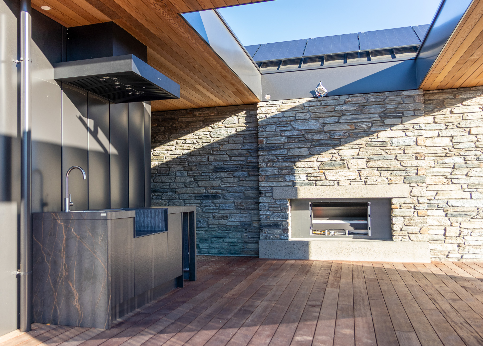 Modern outdoor kitchen with a dark marble countertop, stainless steel sink, extractor hood, and a stone wall with built-in fireplace under a partly open wooden ceiling.