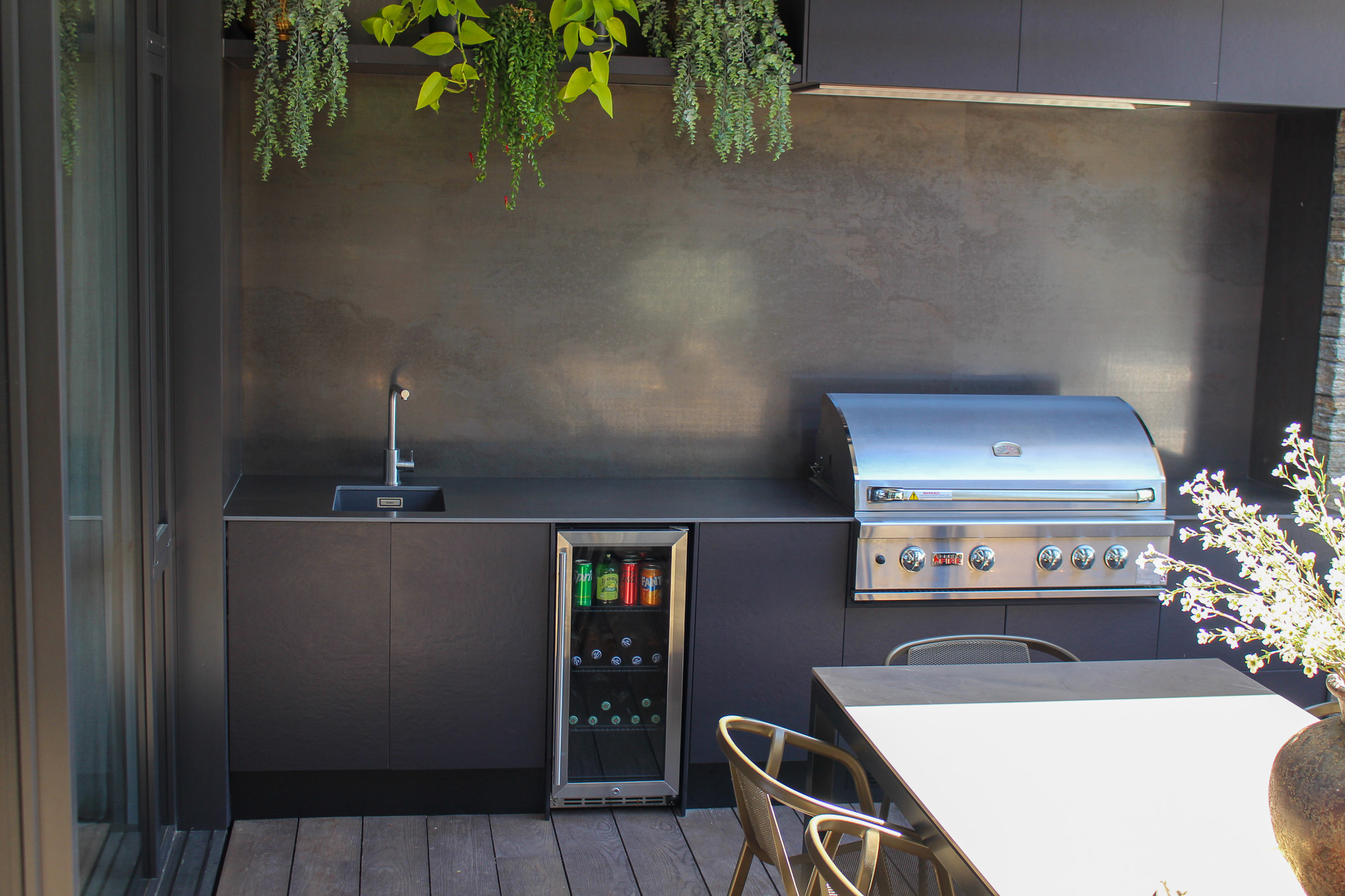 Modern outdoor kitchen with black countertops, a built-in stainless steel grill, a small sink, a beverage fridge stocked with drinks, and a dining table with metal chairs.