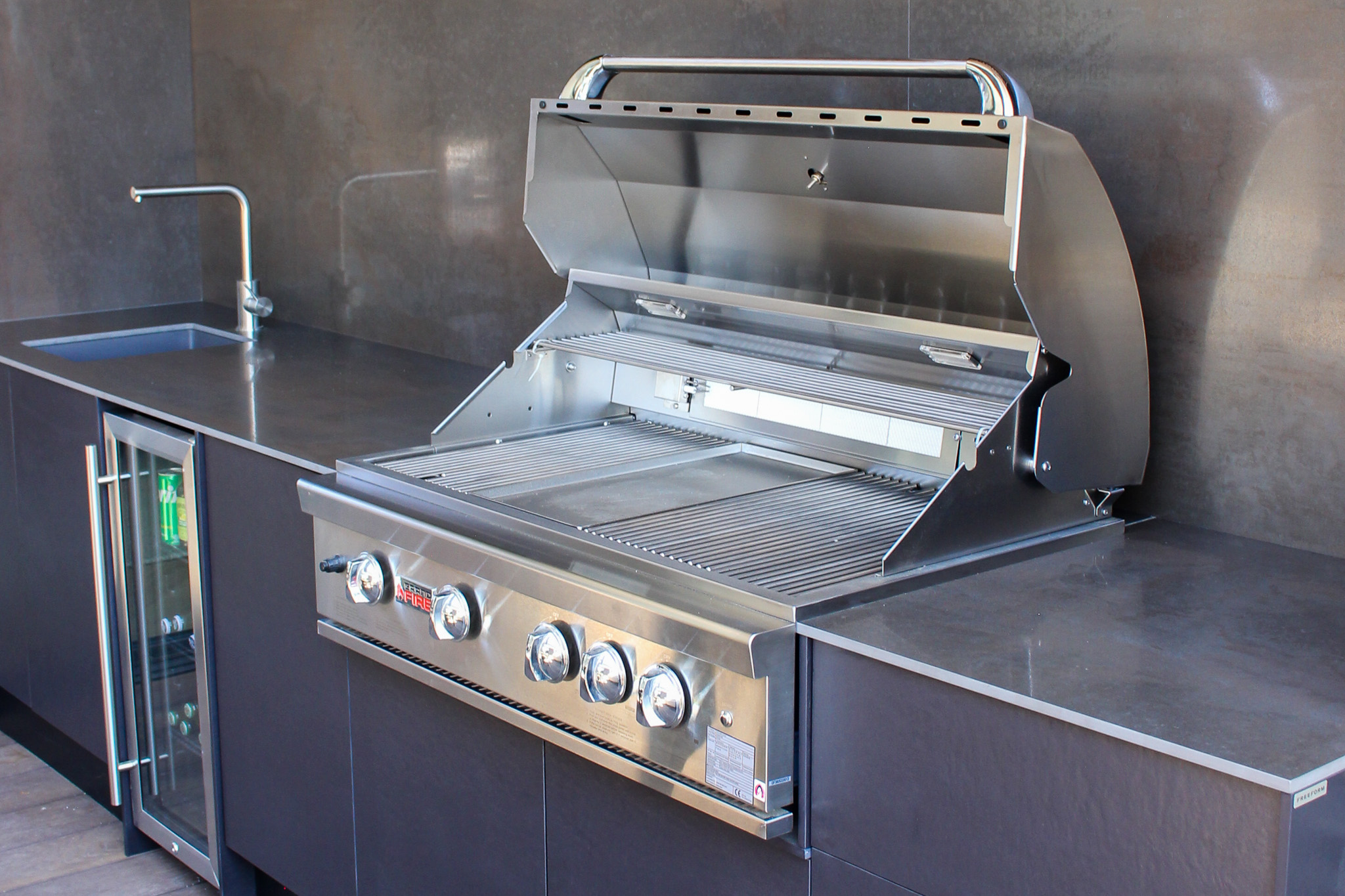 Modern outdoor kitchen with a stainless steel gas barbecue grill, a sink, and a beverage fridge under dark countertops.