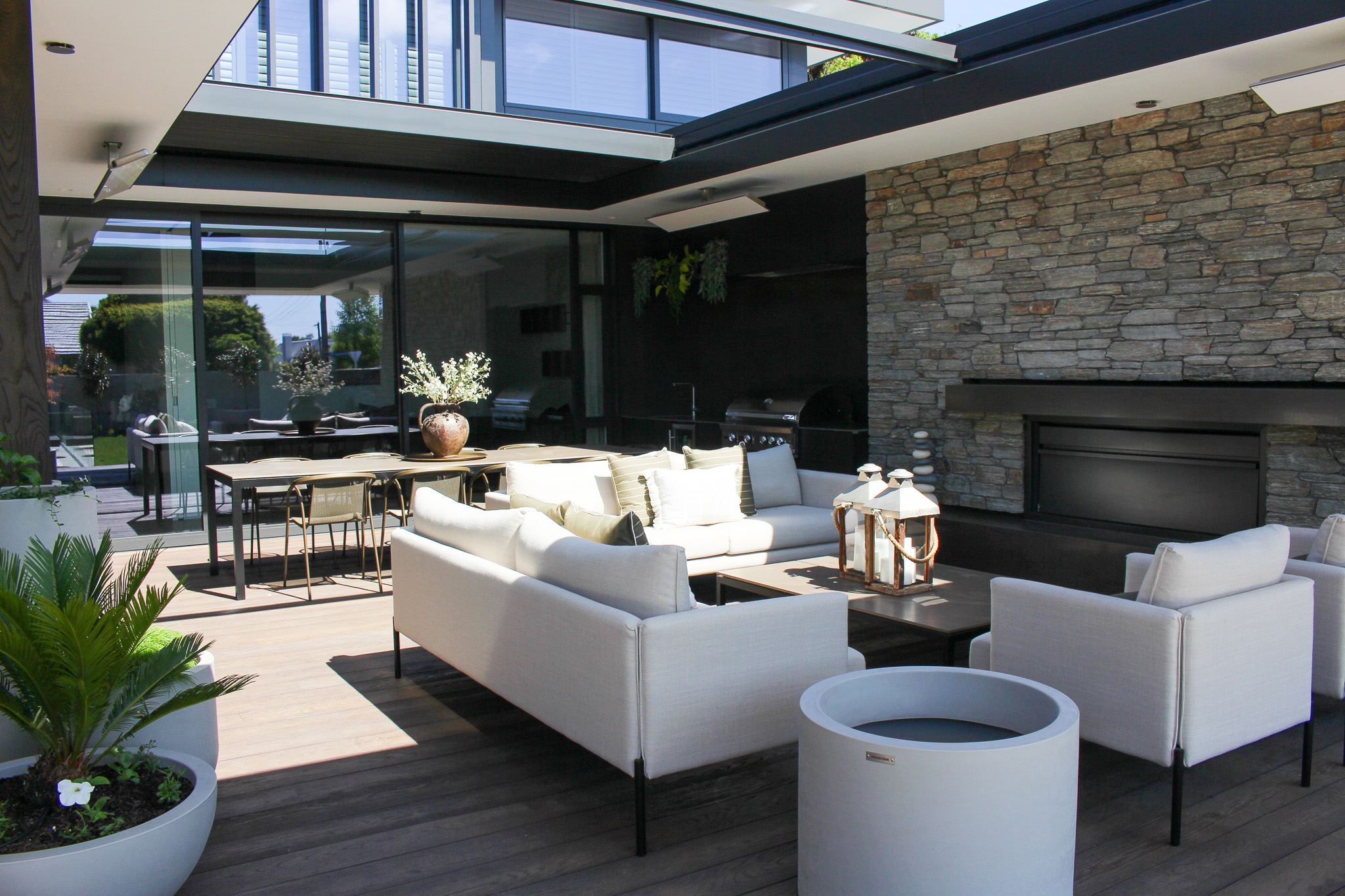 Modern outdoor patio with beige sofas and armchairs around a coffee table, a stone wall with a fireplace, and a dining table with chairs in the background.