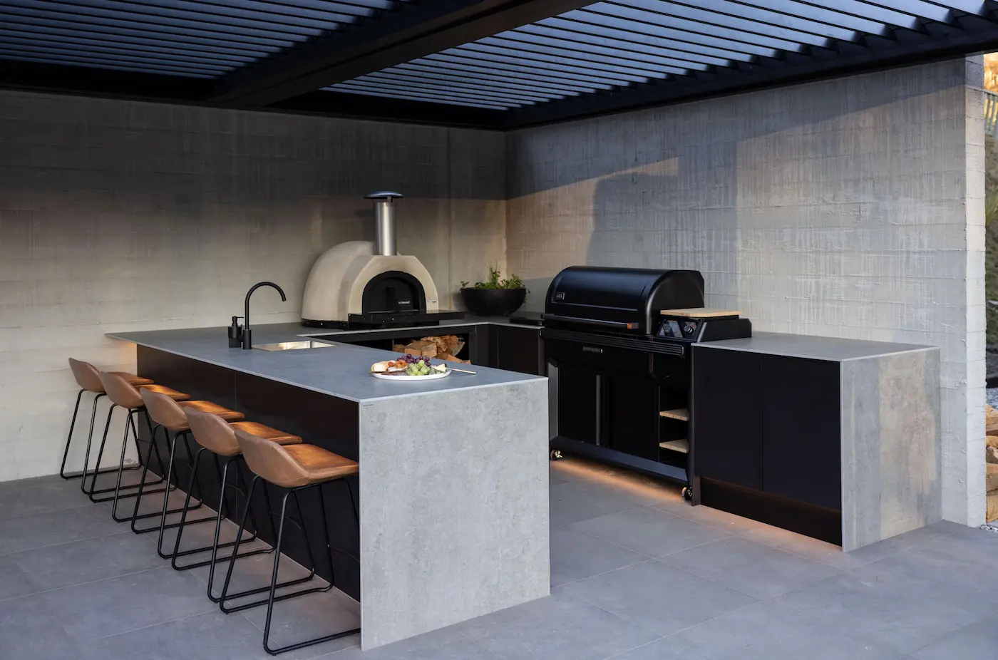 Modern outdoor kitchen with a concrete island, brown bar stools, built-in pizza oven, grill, and under-counter storage.