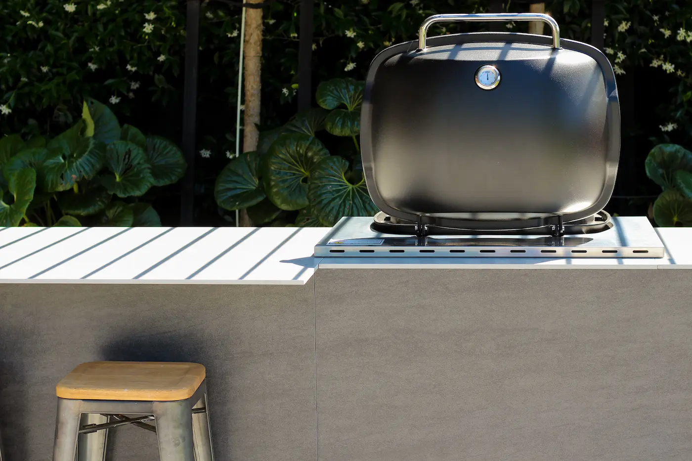 Modern outdoor kitchen with a inbuilt weber grill and a metal stool with a wooden seat, set against green foliage.