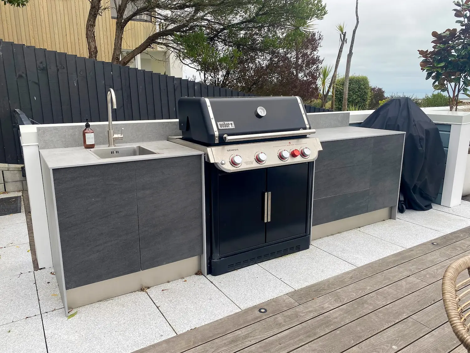 Modern outdoor kitchen with a stainless steel sink, Weber Genesis gas grill, and grey cabinetry on a tiled patio.
