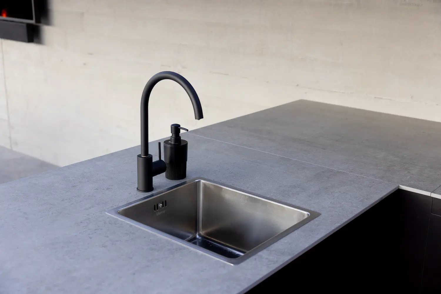 Modern outdoor kitchen sink with a matte black curved faucet and soap dispenser set in a grey countertop.