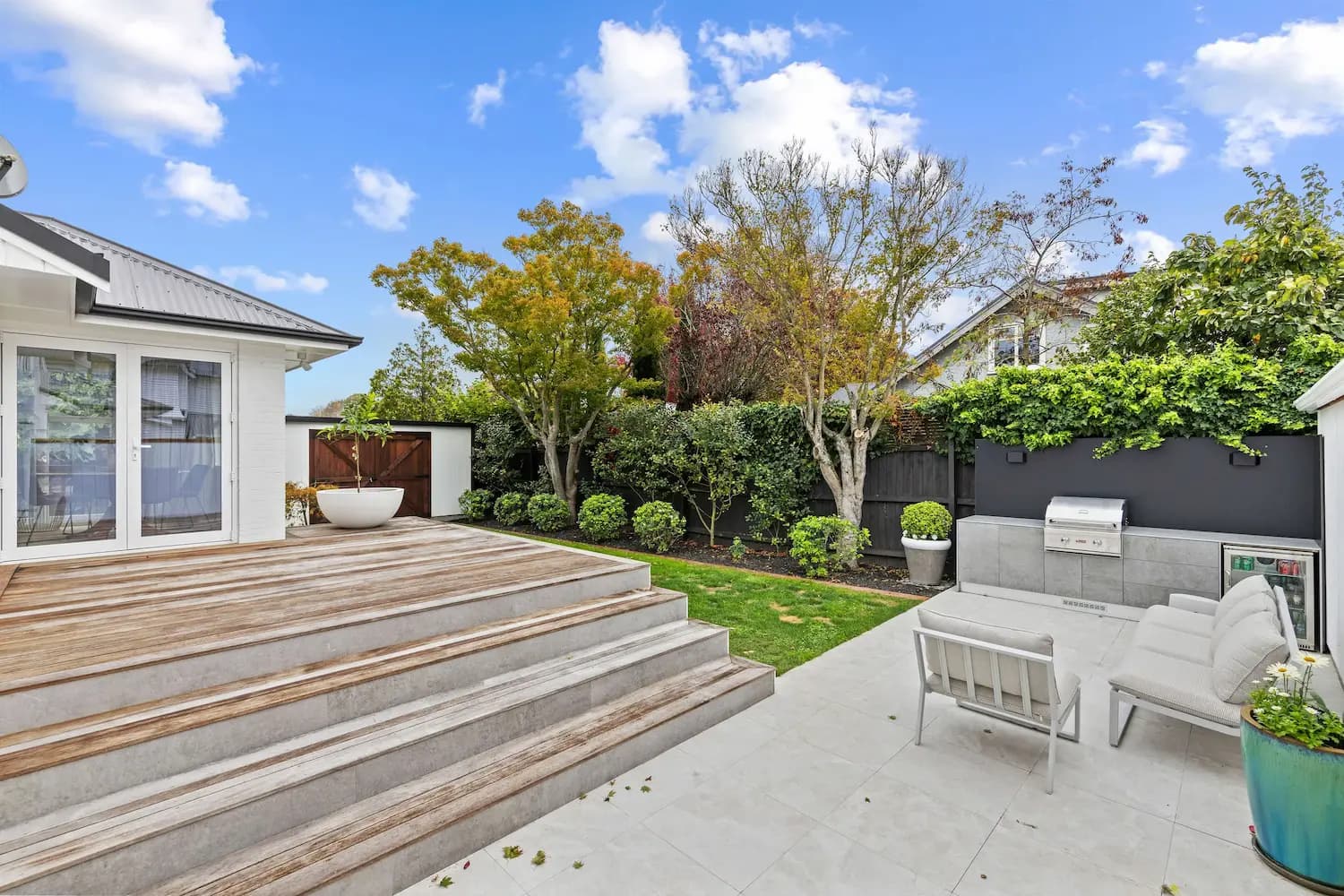 Outdoor kitchen with wooden steps, white cushioned seating, built-in barbecue, and greenery under a blue sky.
