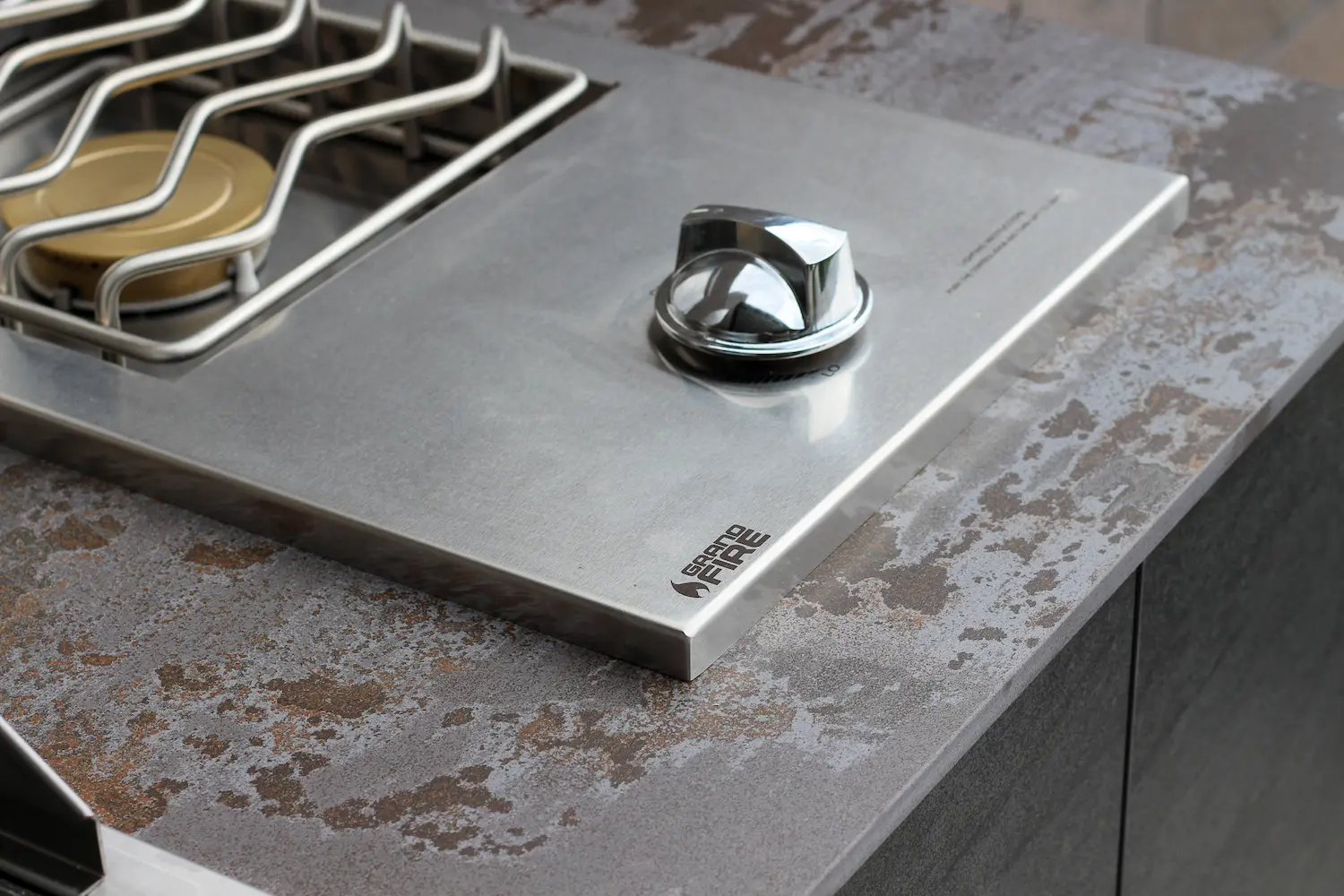 Close-up of a stainless steel Grand Fire gas burner control knob on a textured outdoor kitchen countertop.