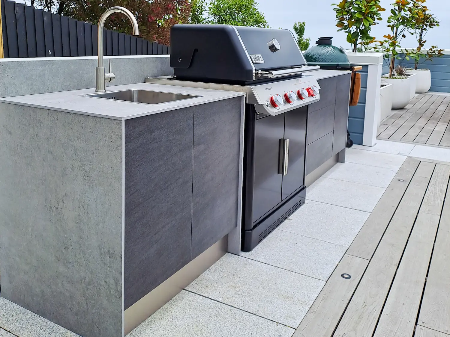 Modern outdoor kitchen with stainless steel sink, black gas grill with red control knobs, and wood decking floor.