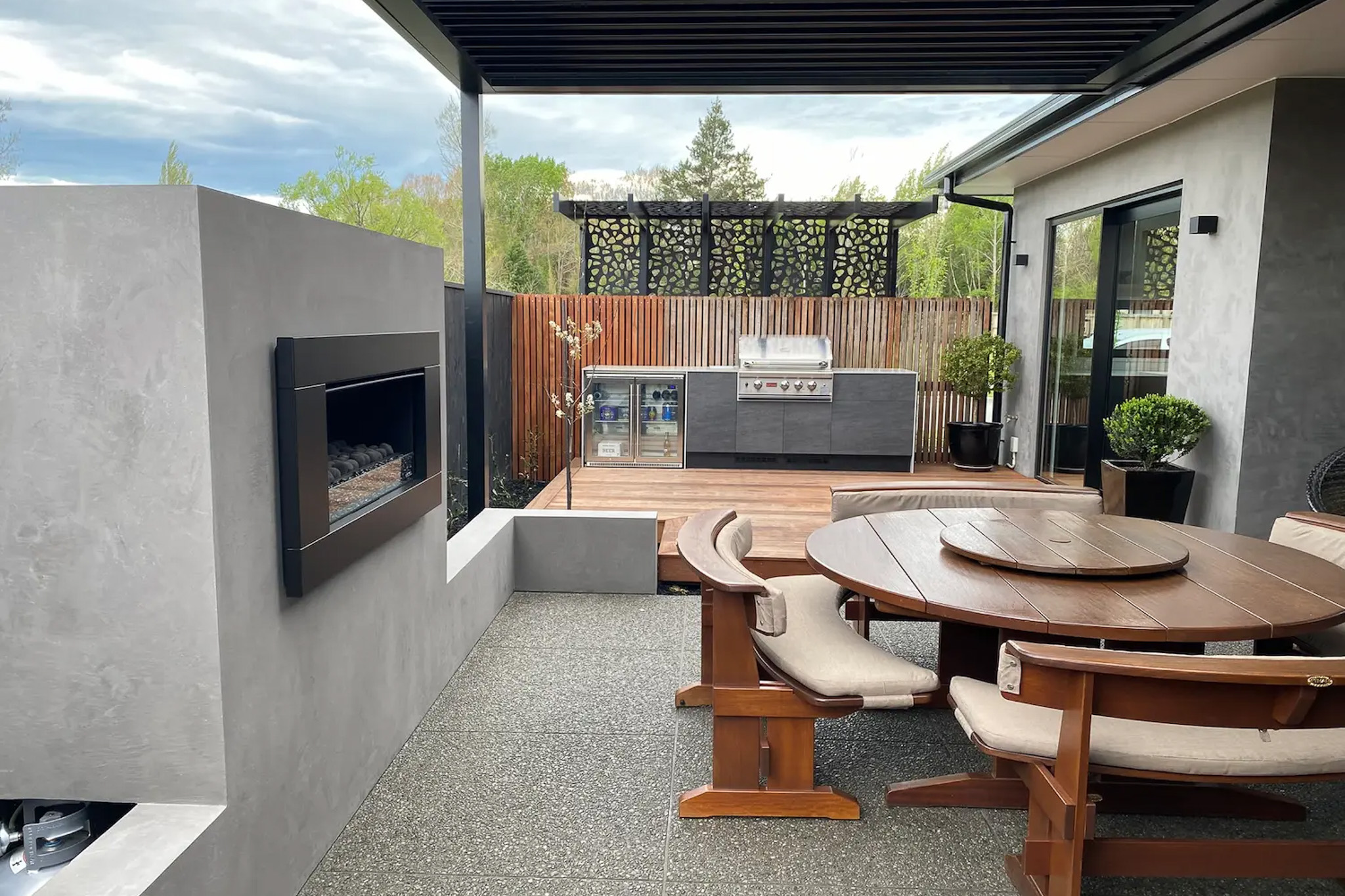 Modern outdoor patio with a built-in gas fireplace, wooden round table with cushioned benches, and a barbecue grill on a wooden deck.