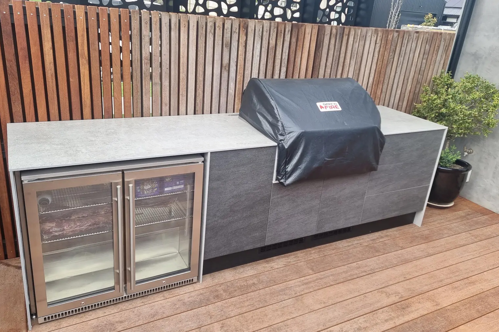 Outdoor kitchen setup with a built-in barbecue covered by a black Grandfire BBQ cover and a double glass-door fridge on a wooden deck.