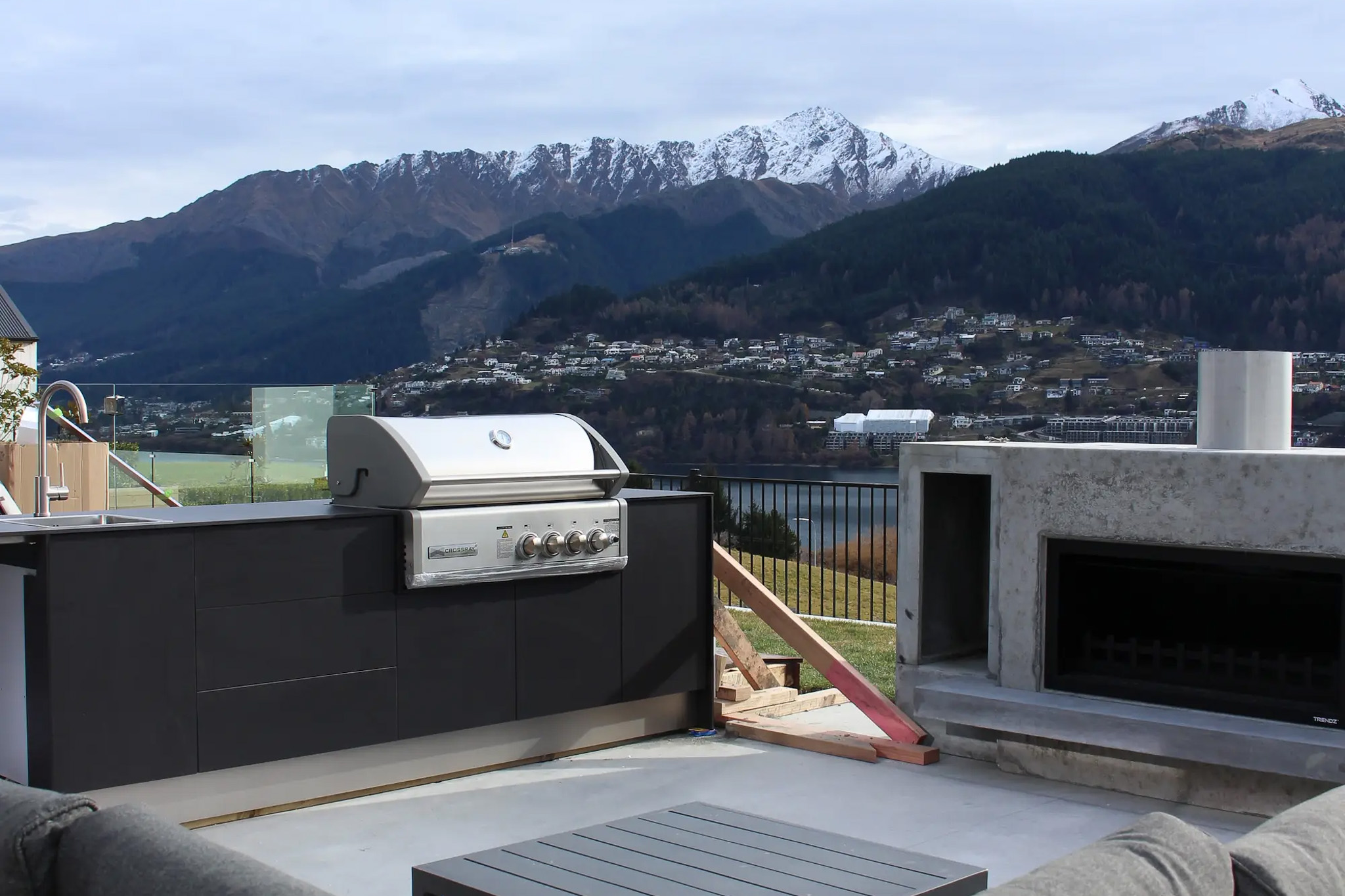 Outdoor kitchen with a built-in stainless steel barbecue grill and a concrete fireplace, overlooking a lake with snow-capped mountains and houses on hills in the background.