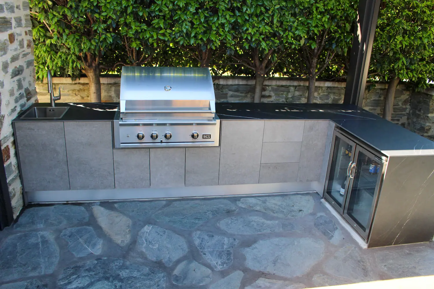 Modern outdoor kitchen with built-in stainless steel grill, sink, beverage cooler, and dark countertops surrounded by greenery.