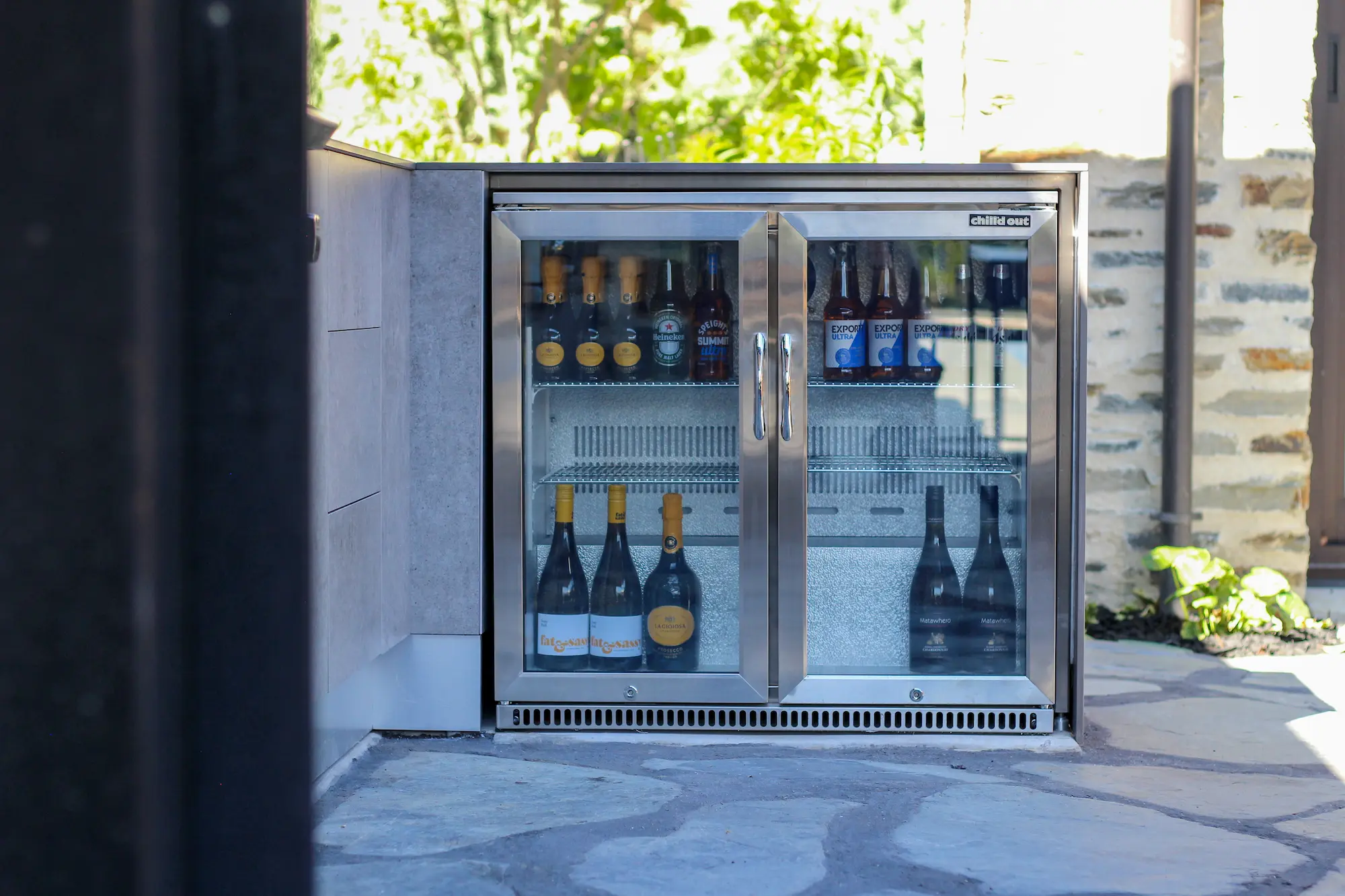 Stainless steel outdoor fridge with glass doors containing various bottled beers and wines.