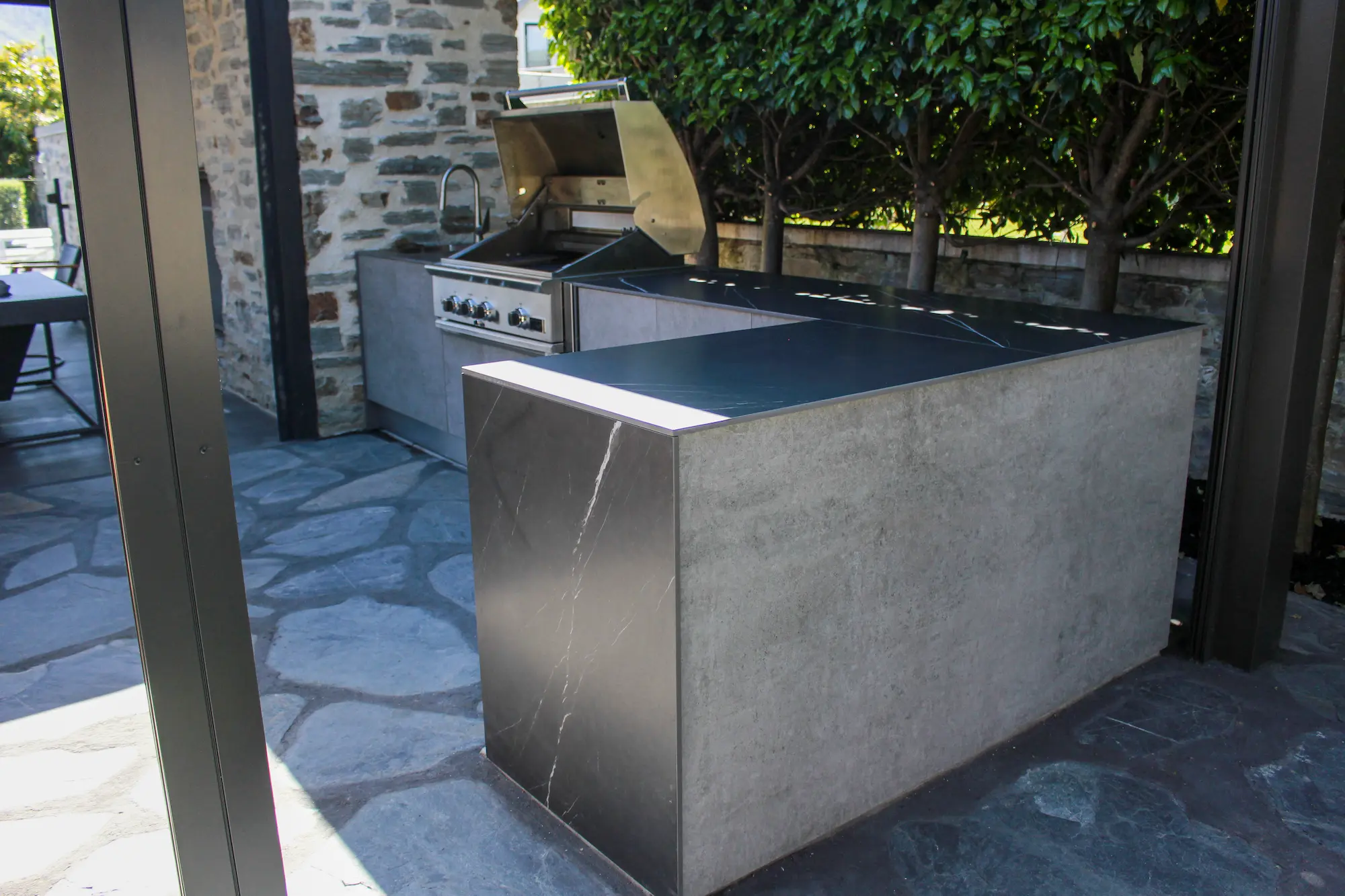 Modern outdoor kitchen with built-in stainless steel gas grill, sink, and stone countertops surrounded by stone walls and greenery.