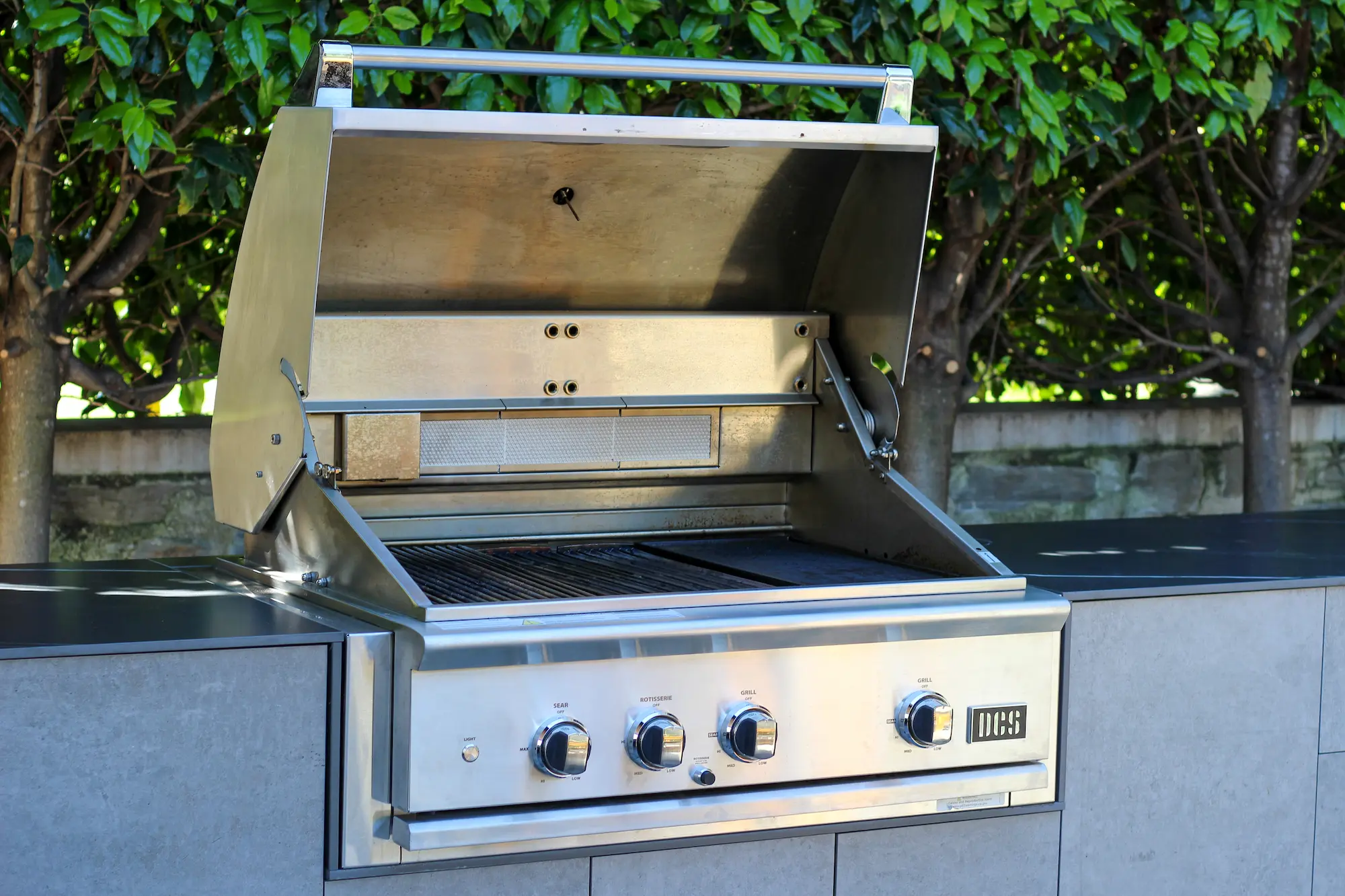 Outdoor stainless steel BBQ grill with open lid and control knobs on grey stone countertop.