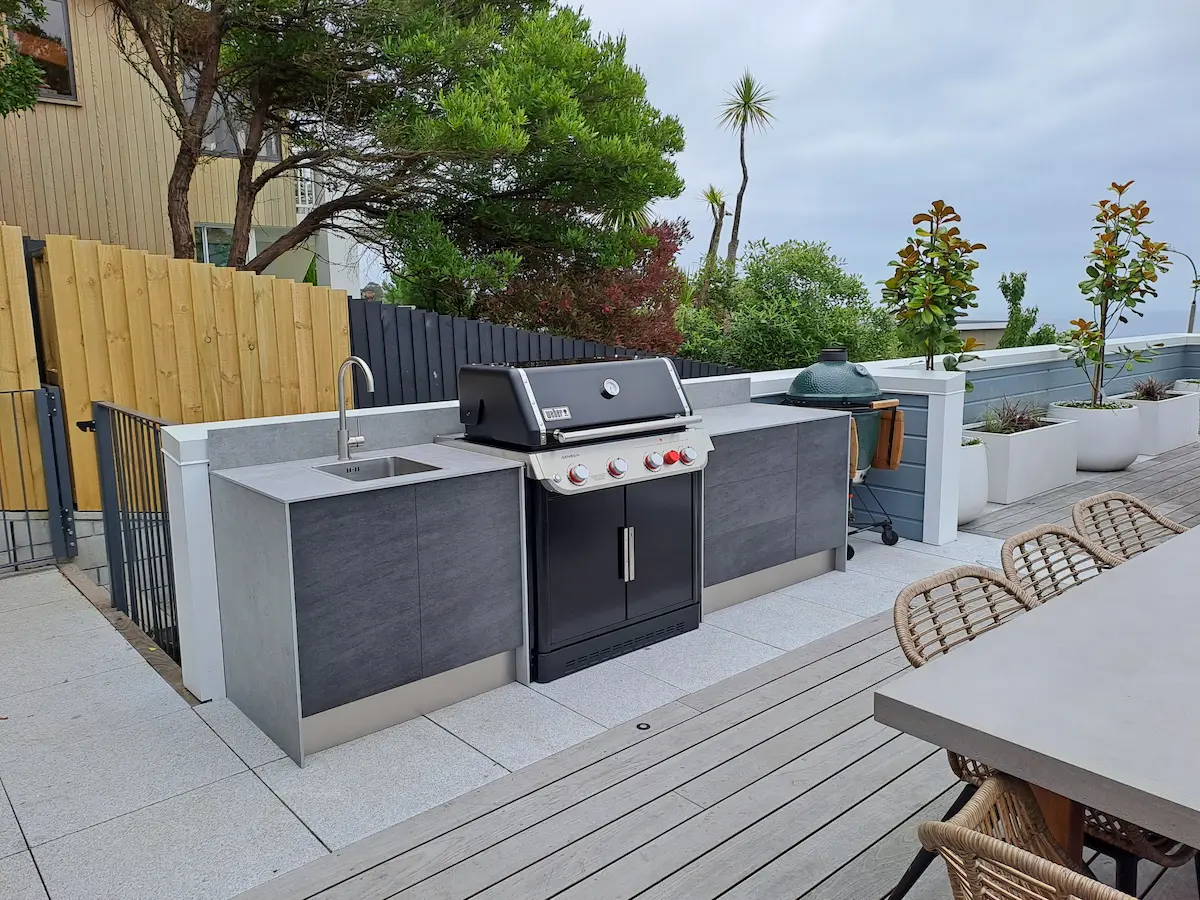 Modern outdoor kitchen with a built-in gas grill, sink, and stone countertops on a tiled patio with wicker chairs and potted plants.