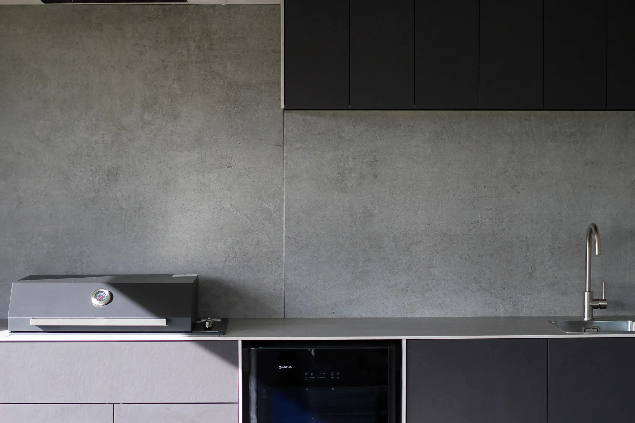 Modern Dekton stone kitchen counter with a built-in grill, black fridge, stainless steel sink, and grey textured backsplash.