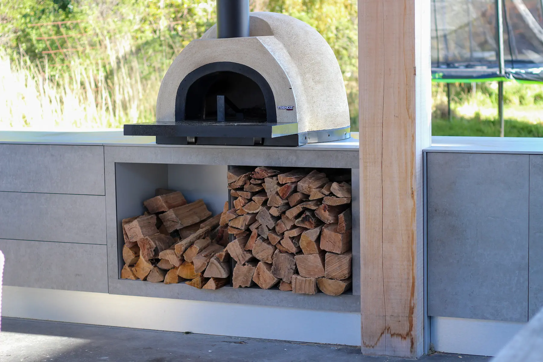 Outdoor kitchen with pizza oven, gas grill, mini fridge, and wicker seating with cushions under a wooden pergola.