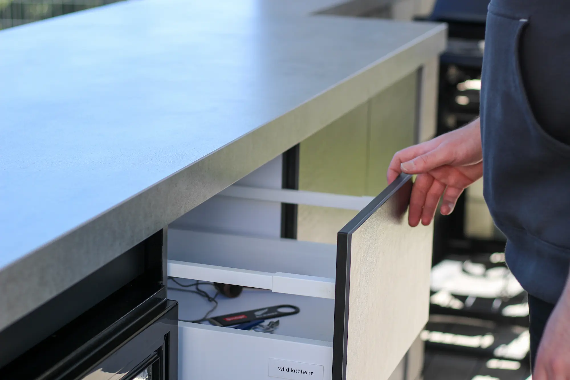 Person opening a kitchen drawer labelled 'wild kitchens' with utensils inside below a grey countertop.