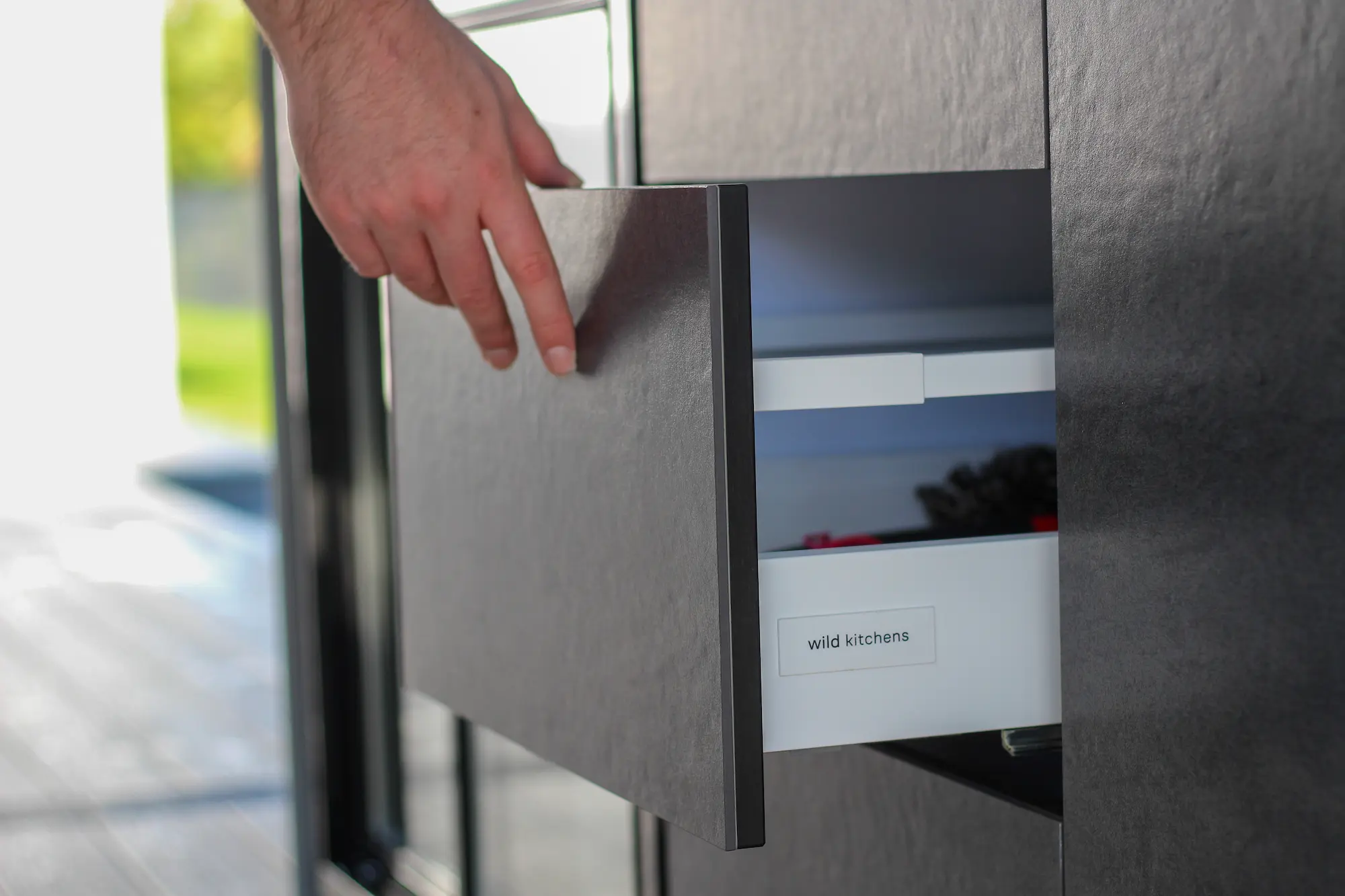 Hand opening a sleek dark grey stone kitchen drawer with 'wild kitchens' label inside.