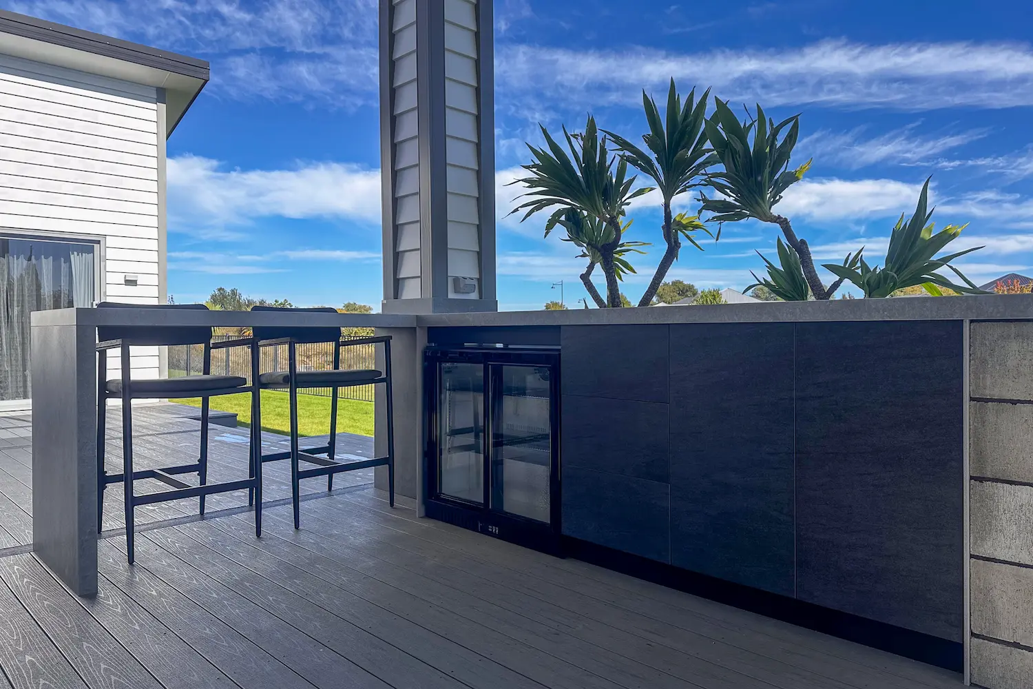 Outdoor patio area with modern bar counter, two black bar stools, mini fridge, and green plants under a blue sky.
