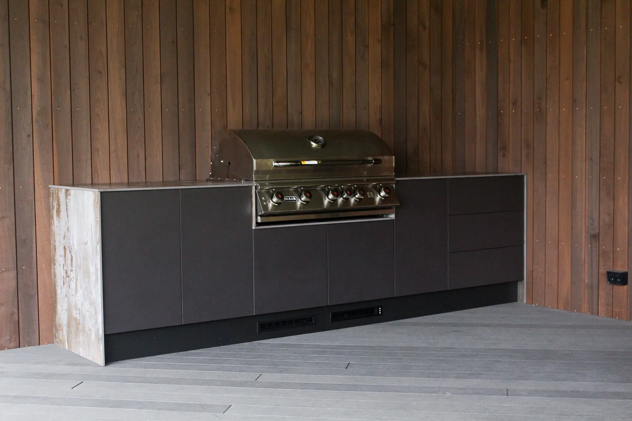 Modern stainless steel outdoor grill integrated into black cabinetry against a wooden panel wall.