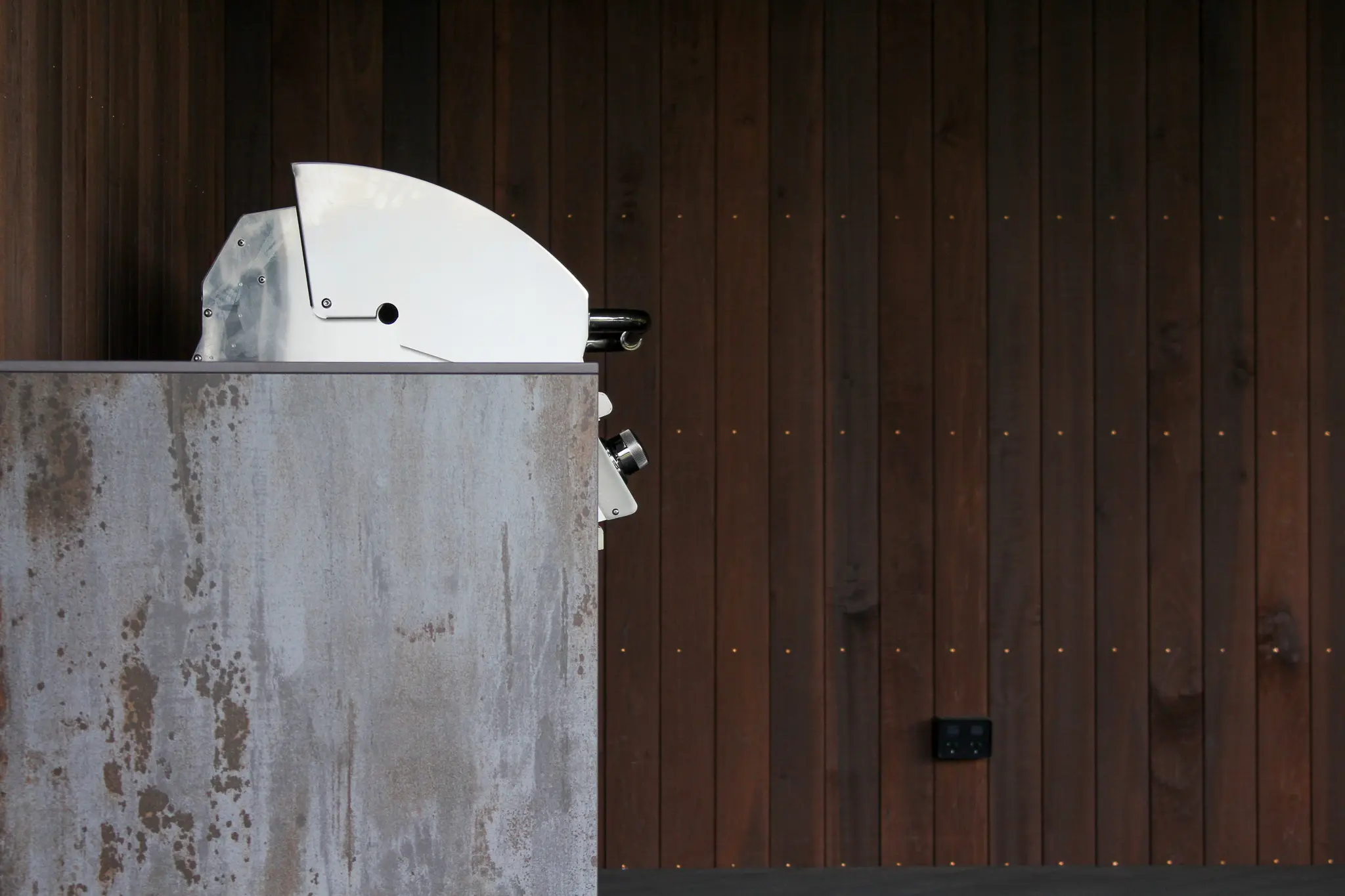 Silver industrial machine partially visible behind a weathered metal panel with a dark wood plank wall background.