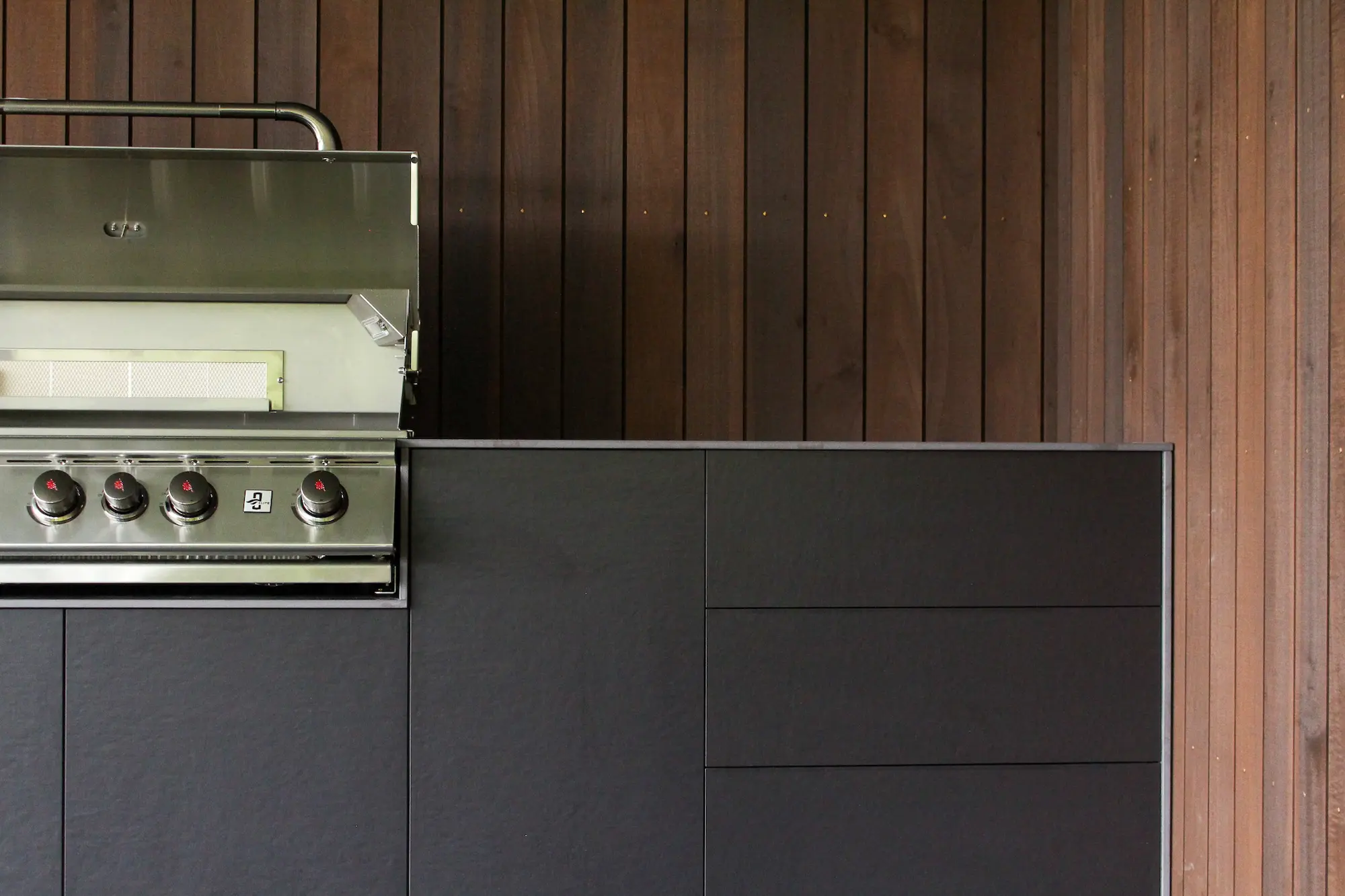 Modern outdoor bbq with four control knobs set into a sleek black cabinet against a wooden panel wall.
