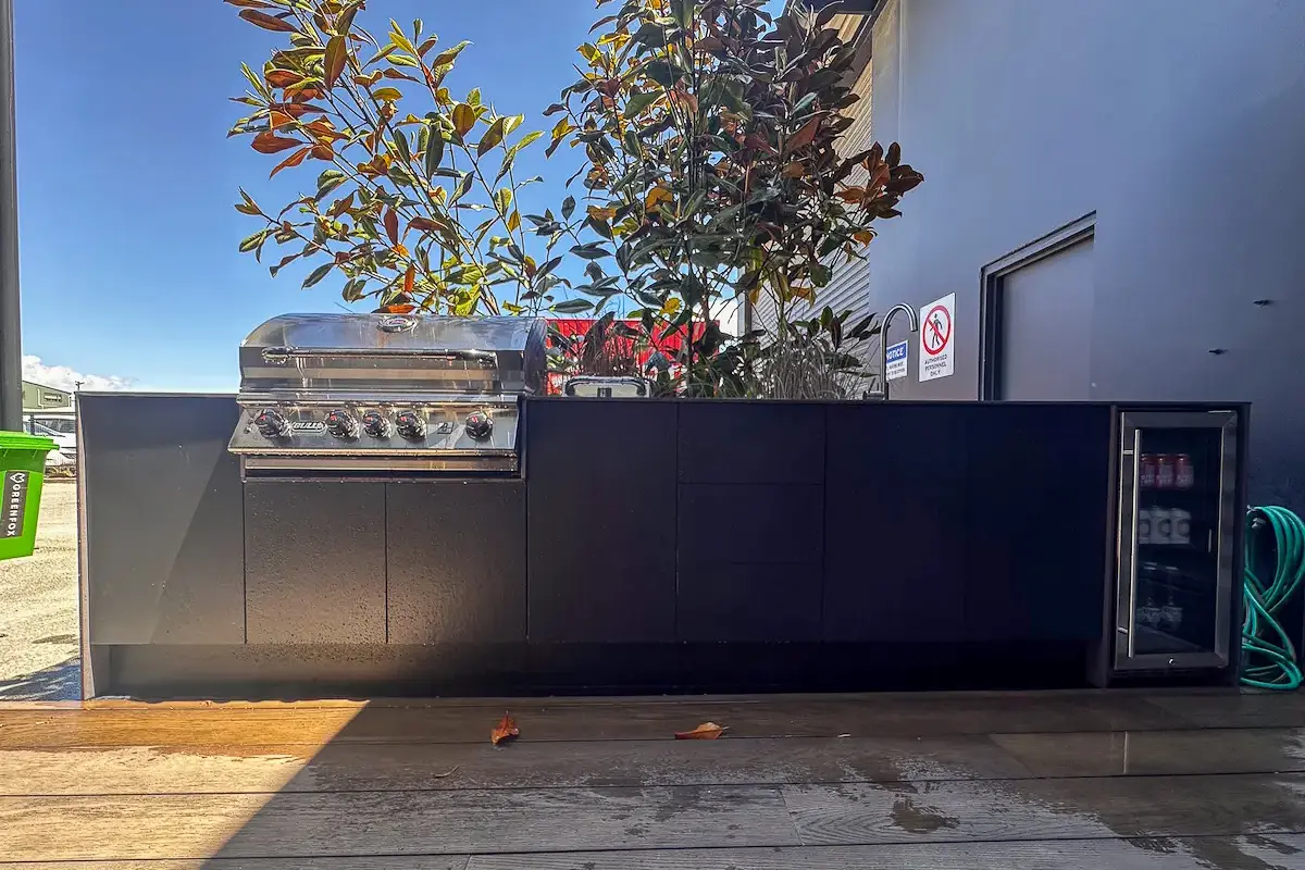 Stone Outdoor kitchen unit with a stainless steel gas grill on the left, a sink in the middle, and a glass-door beverage fridge on the right under a clear blue sky.