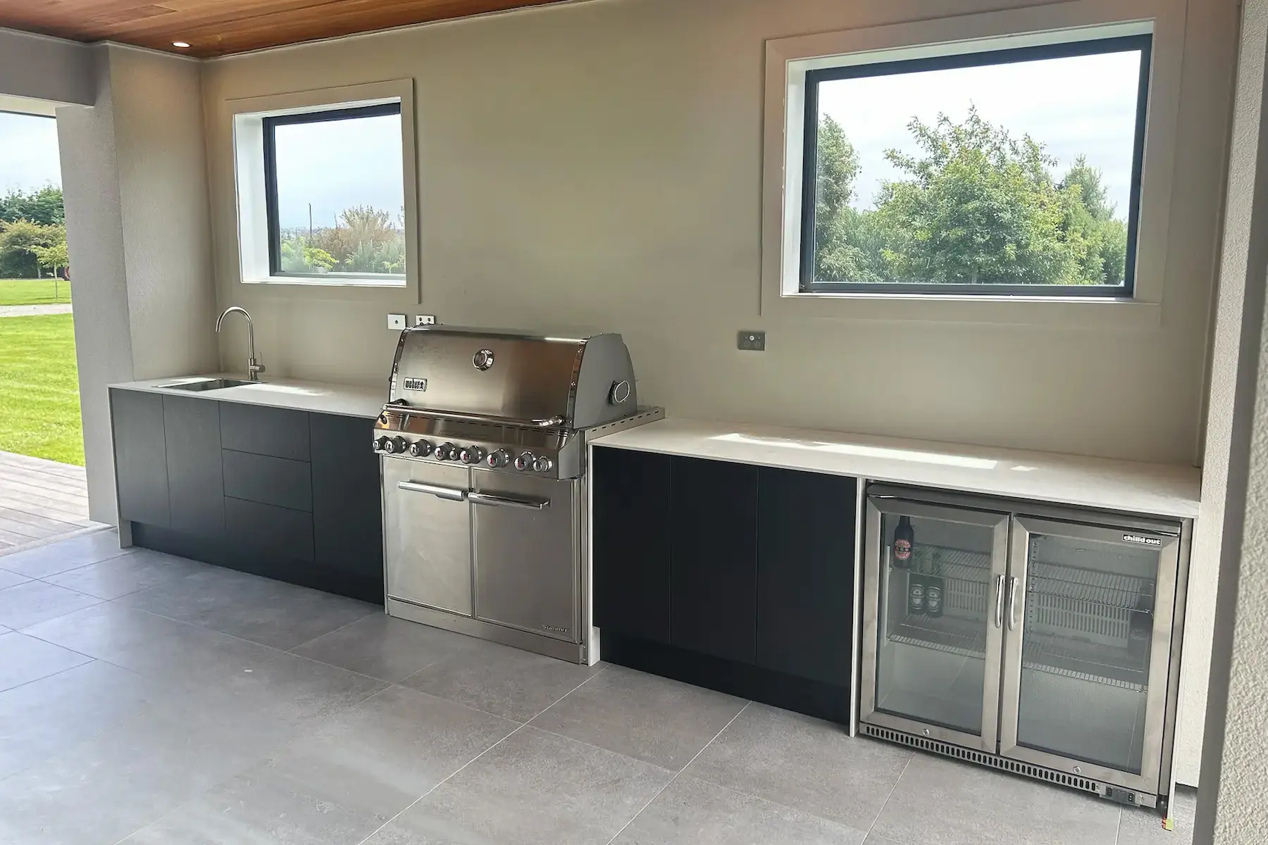 Modern outdoor kitchen with stainless steel barbecue grill, black cabinets, a sink under one window, and a glass-door beverage fridge under another window.