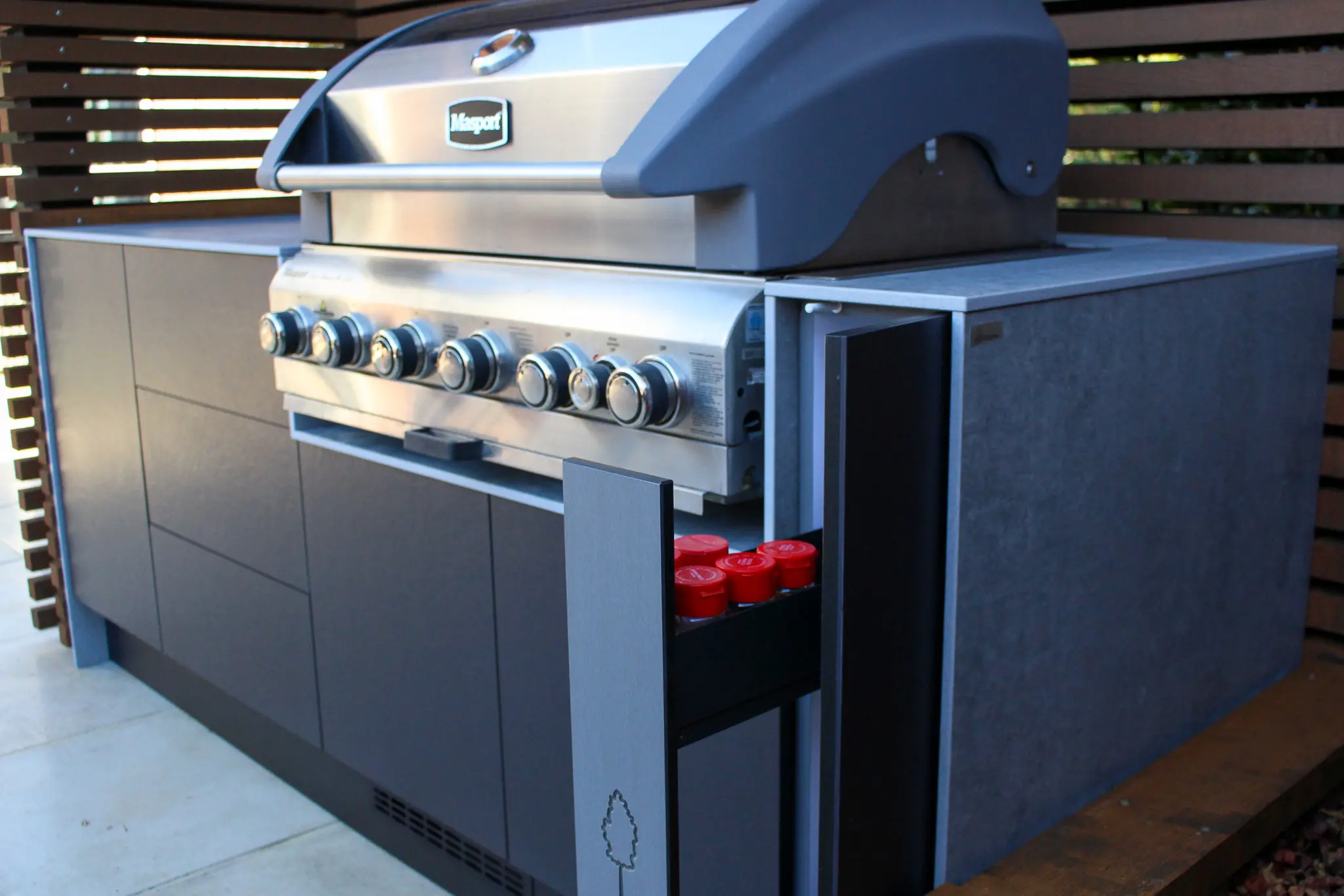 Modern outdoor barbecue grill with six control knobs and a side drawer holding red spice jars.
