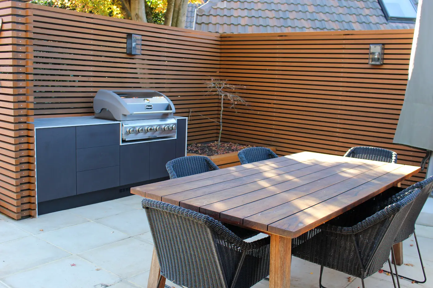 Outdoor kitchen with pizza oven, gas grill, mini fridge, and wicker seating with cushions under a wooden pergola.