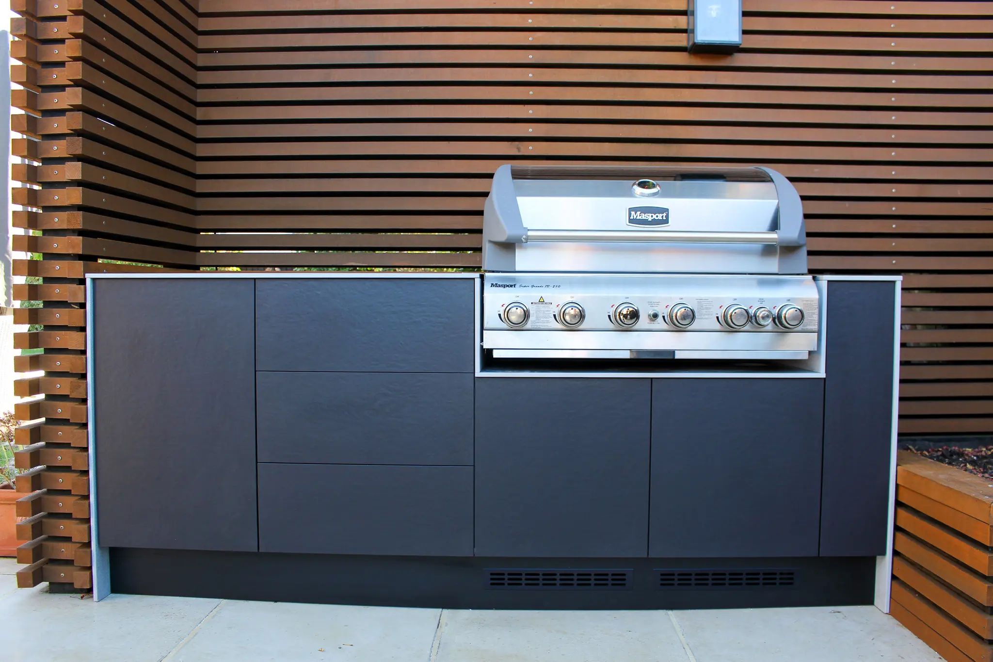 Outdoor built-in Masport stainless steel gas barbecue with black cabinetry against wooden slat wall.