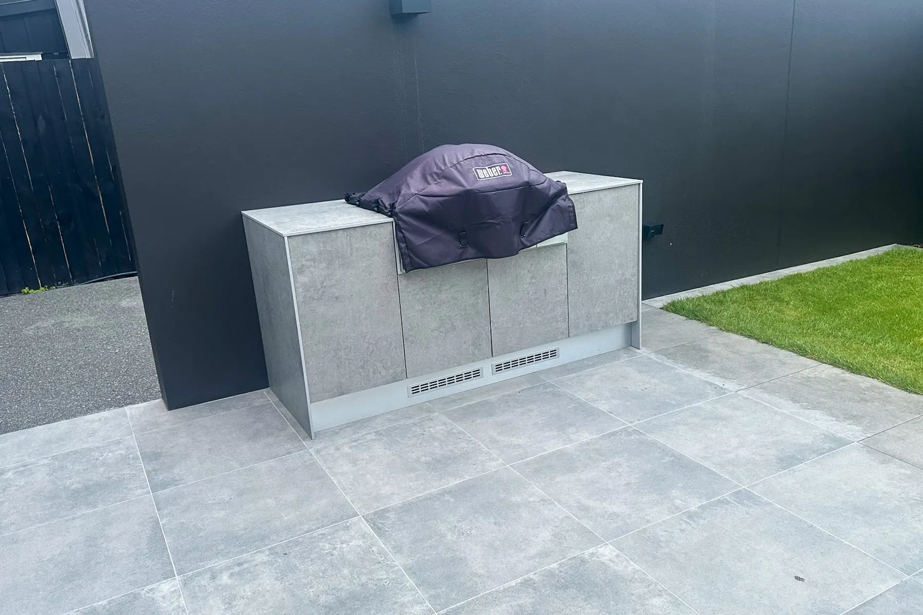 Outdoor kitchen cabinet unit with a covered Weber barbecue against a dark wall on a tiled patio near grass.
