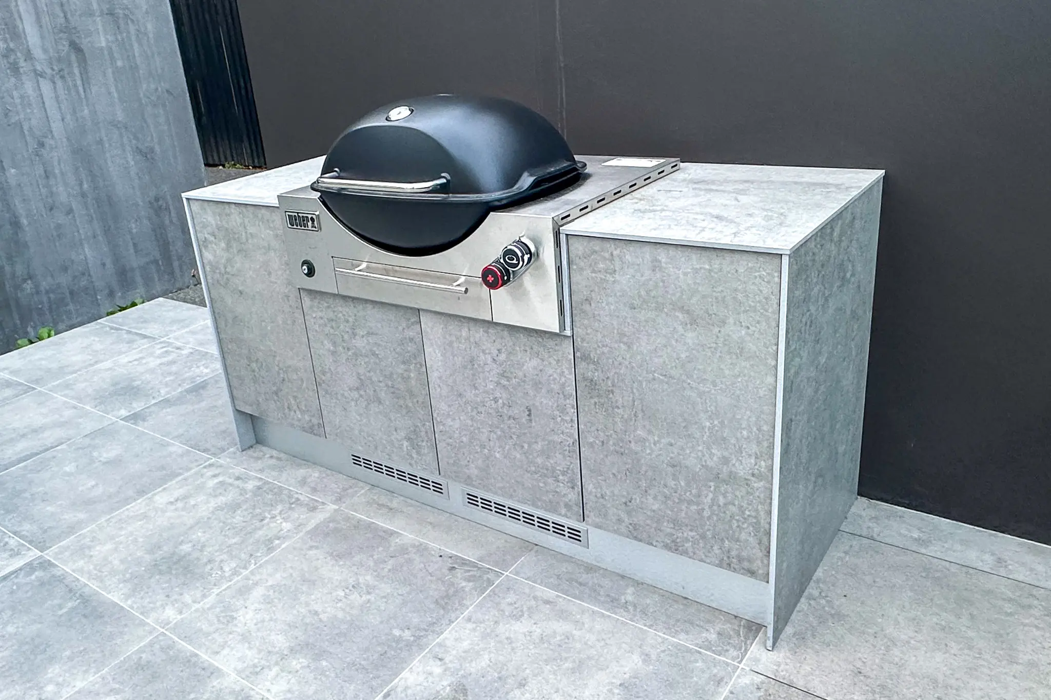 Modern outdoor built-in Weber barbecue grill with grey cabinetry on tiled patio.