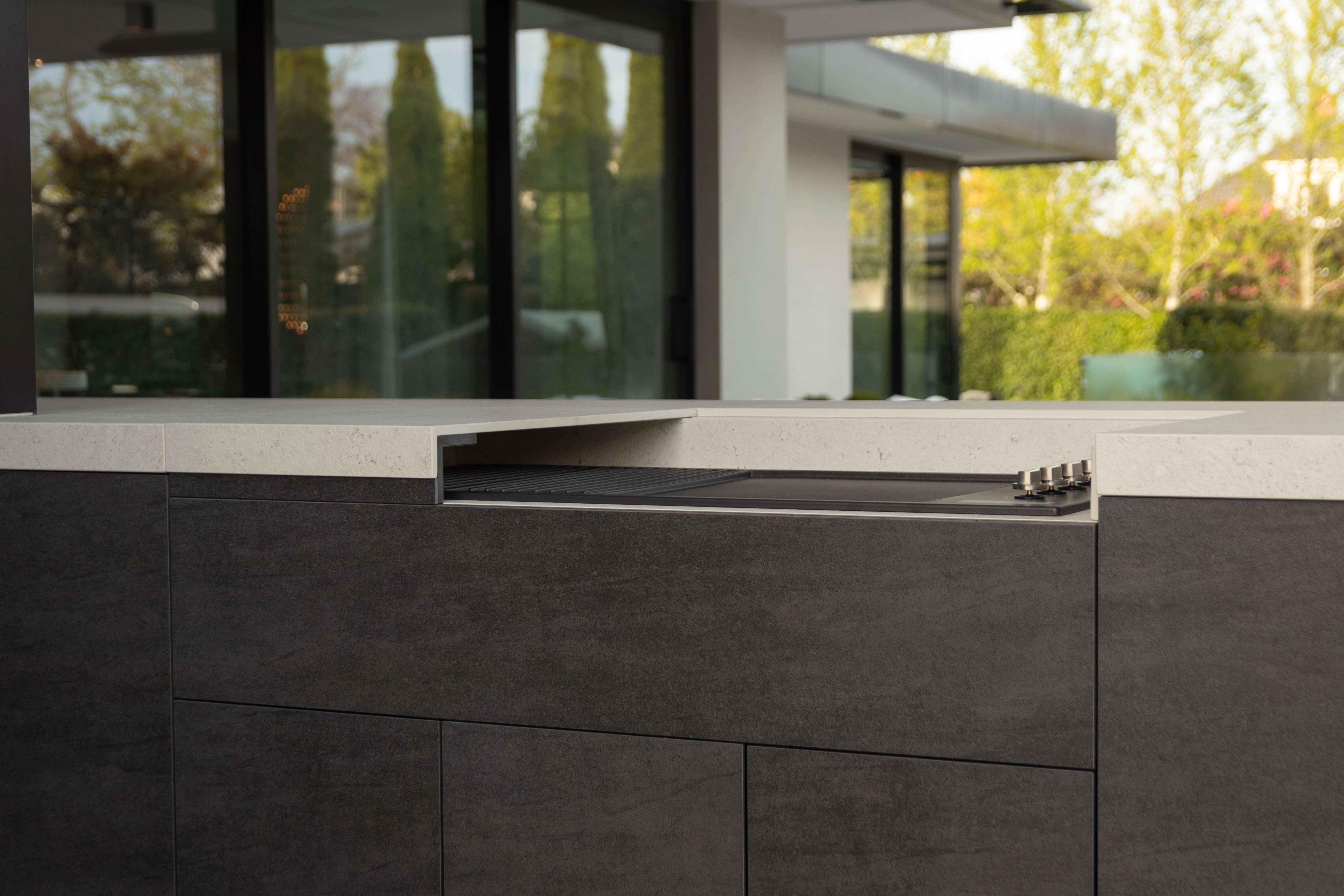 Person reaching up to open a black Stone overhead kitchen cabinet in a modern kitchen with grey and black Stone cabinetry, a small sink, and a built-in barbecue grill.
