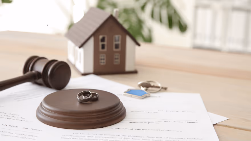 Two wedding rings rest on a judge’s gavel beside legal divorce documents, with a small model house and a set of house keys in the background.