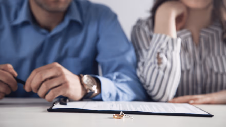 A couple sits at a table with divorce papers in front of them, wedding rings placed on the table.