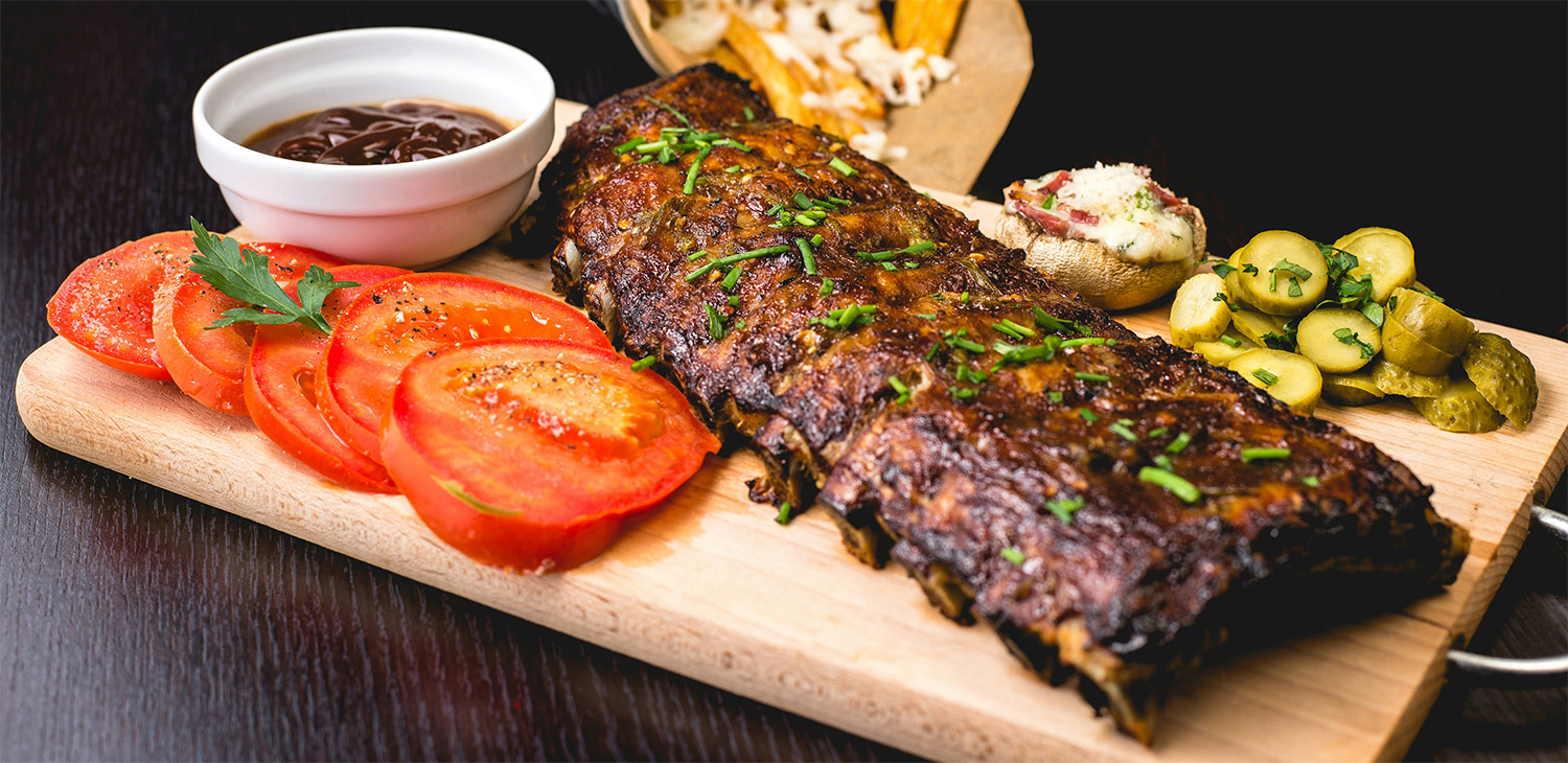 Grilled ribs served on a wooden board with sliced tomatoes, pickles, fries, and barbecue sauce, representing food cost tracking for Dallas restaurants.