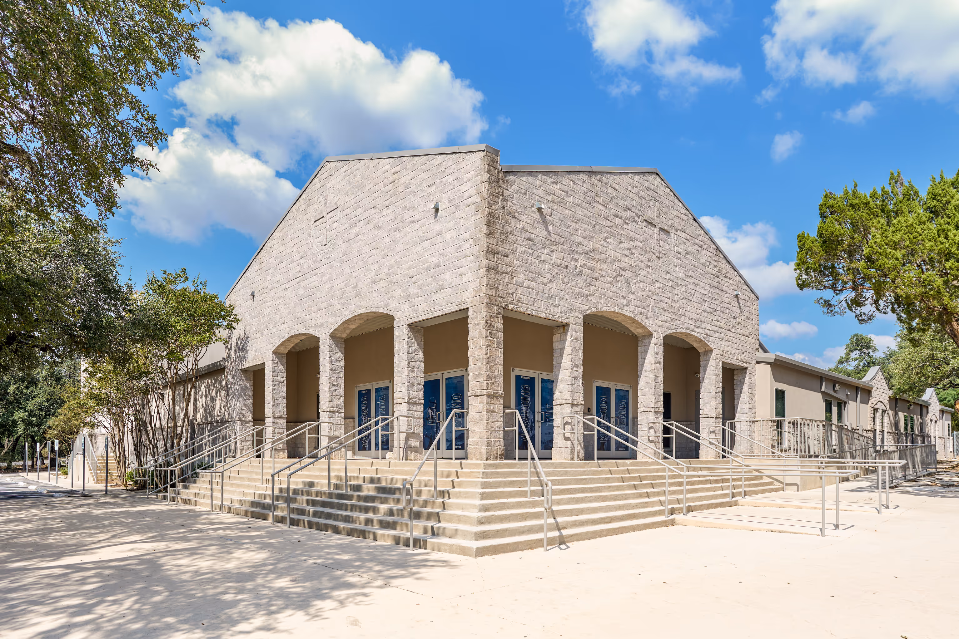 Corner view of a beige stone church building with wide steps, metal railings, and blue entrance doors under arched columns.