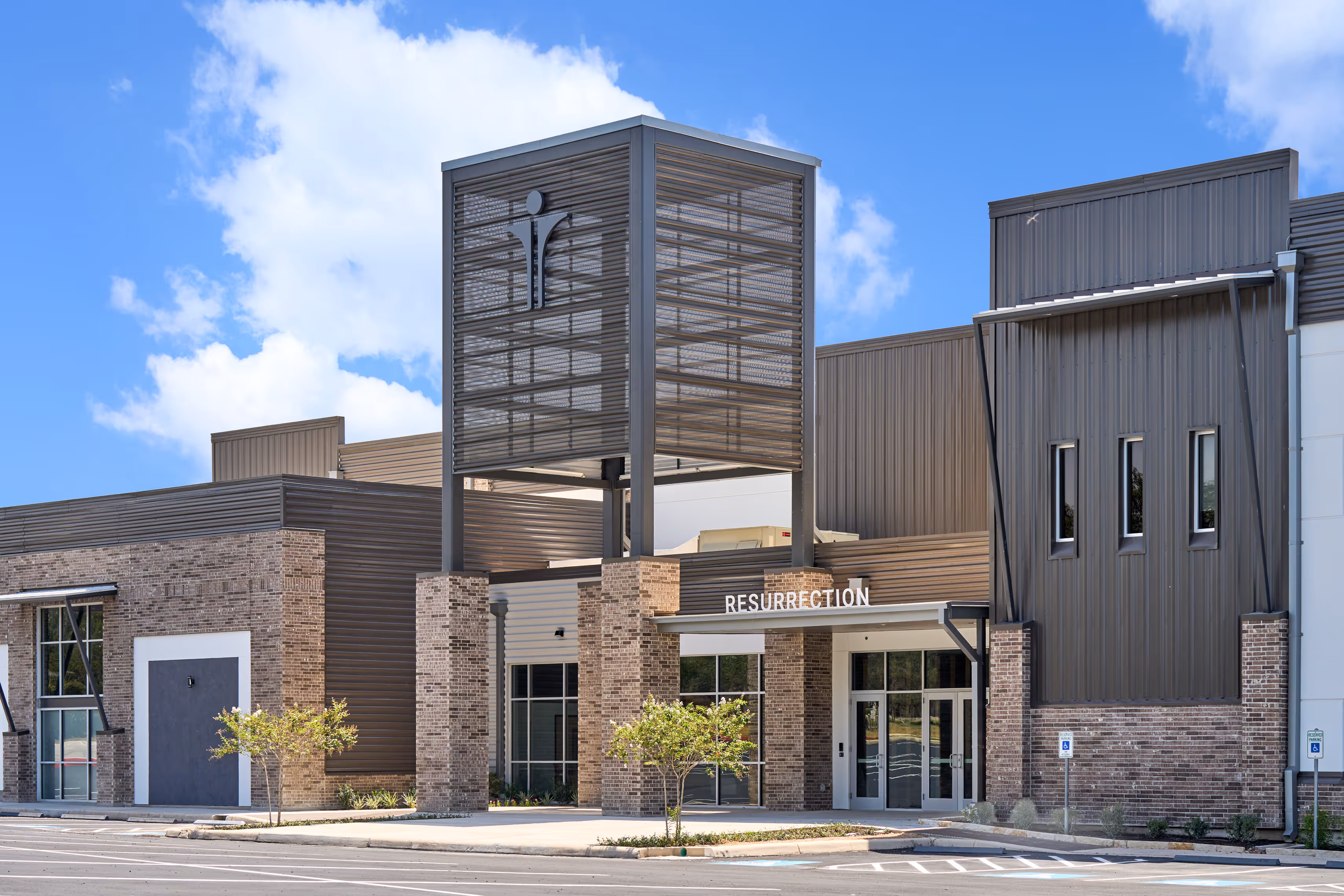 Modern church building with brick and metal exterior, featuring the word 'RESURRECTION' above the entrance and a large metal structure with a cross symbol.