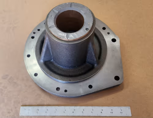 CNC machined cast metal housing with central bore and bolt holes, shown with ruler for scale by Burns Machine Company.