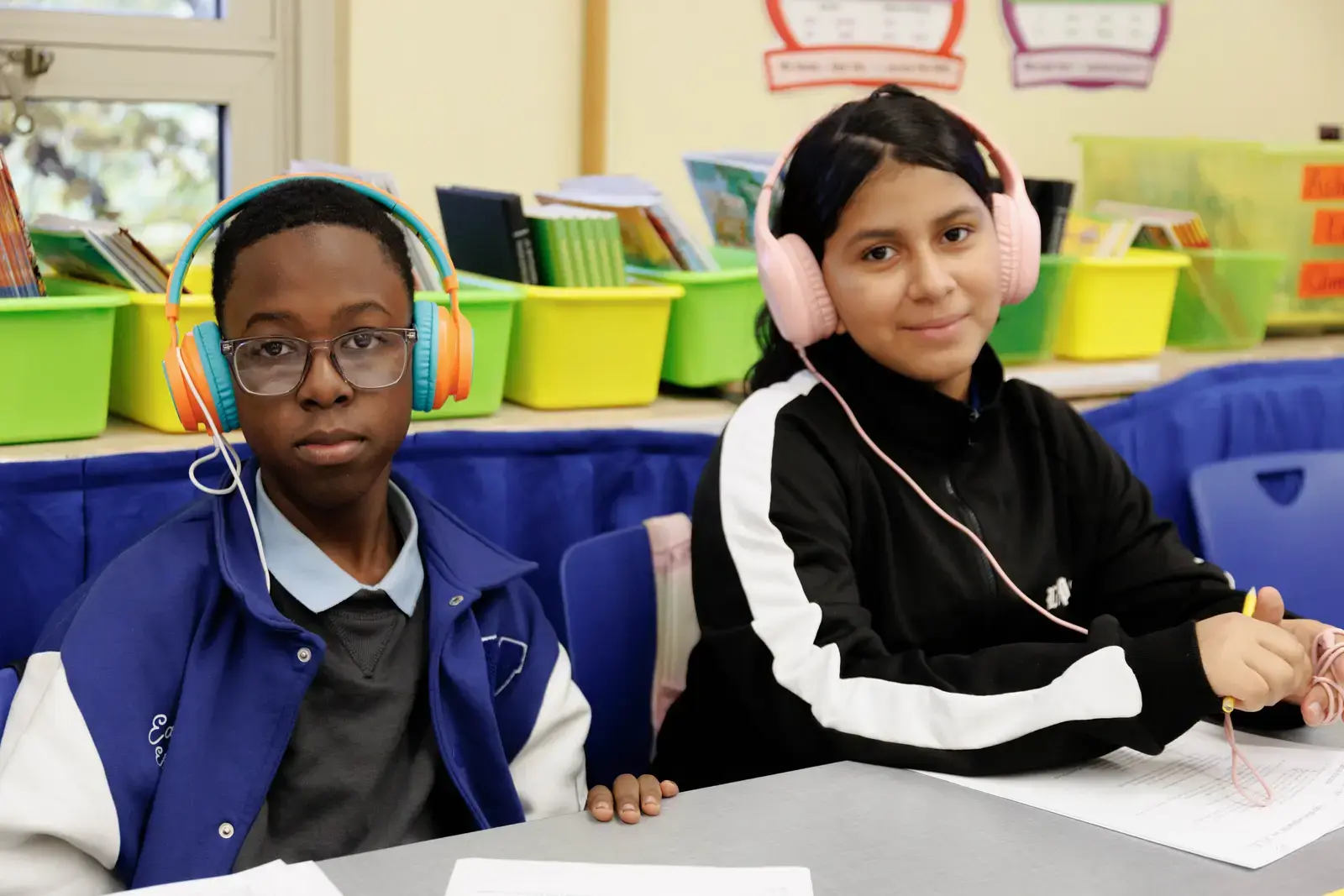 Two students wearing headphones work at a classroom desk