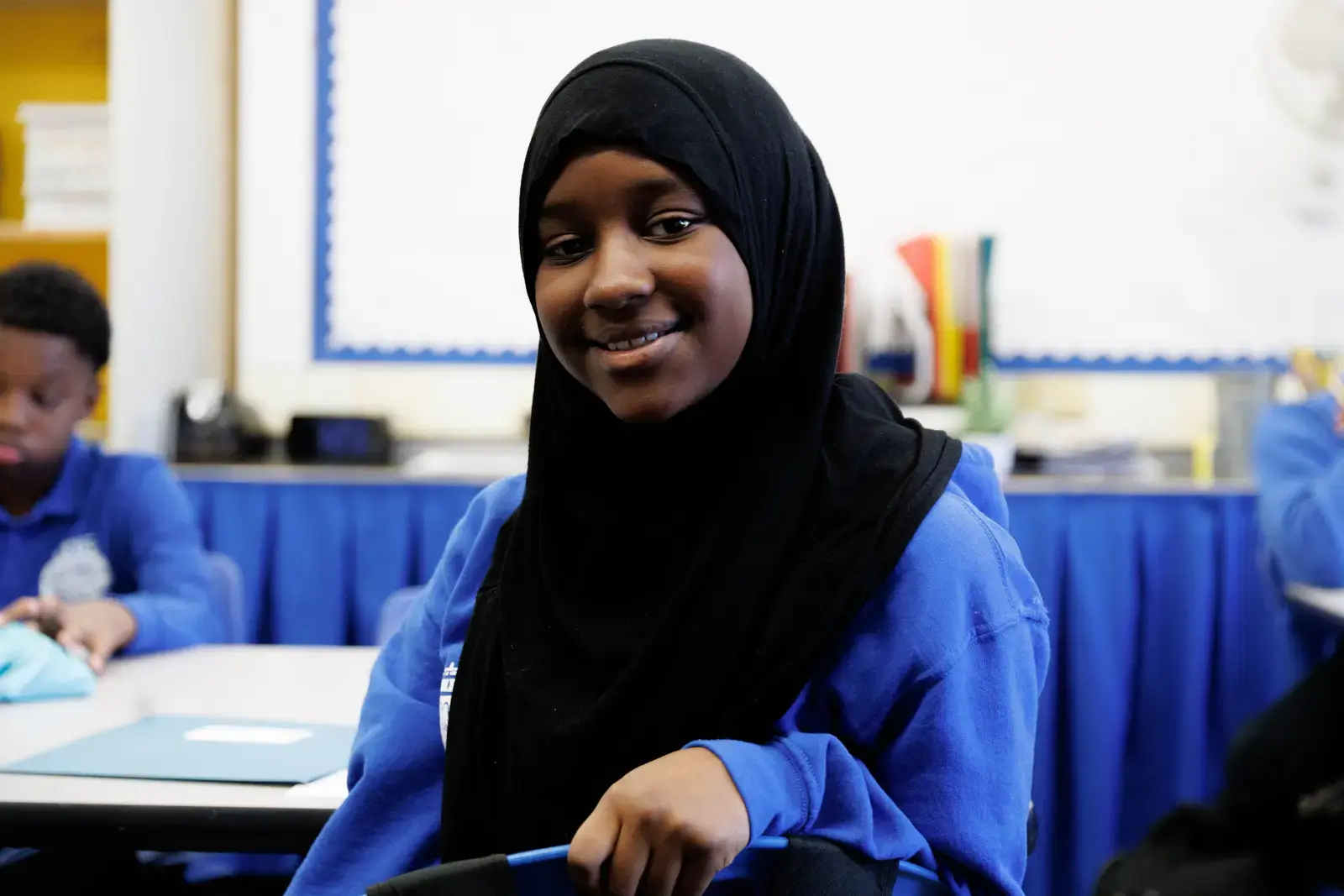Smiling student in hijab and blue uniform seated at classroom desk