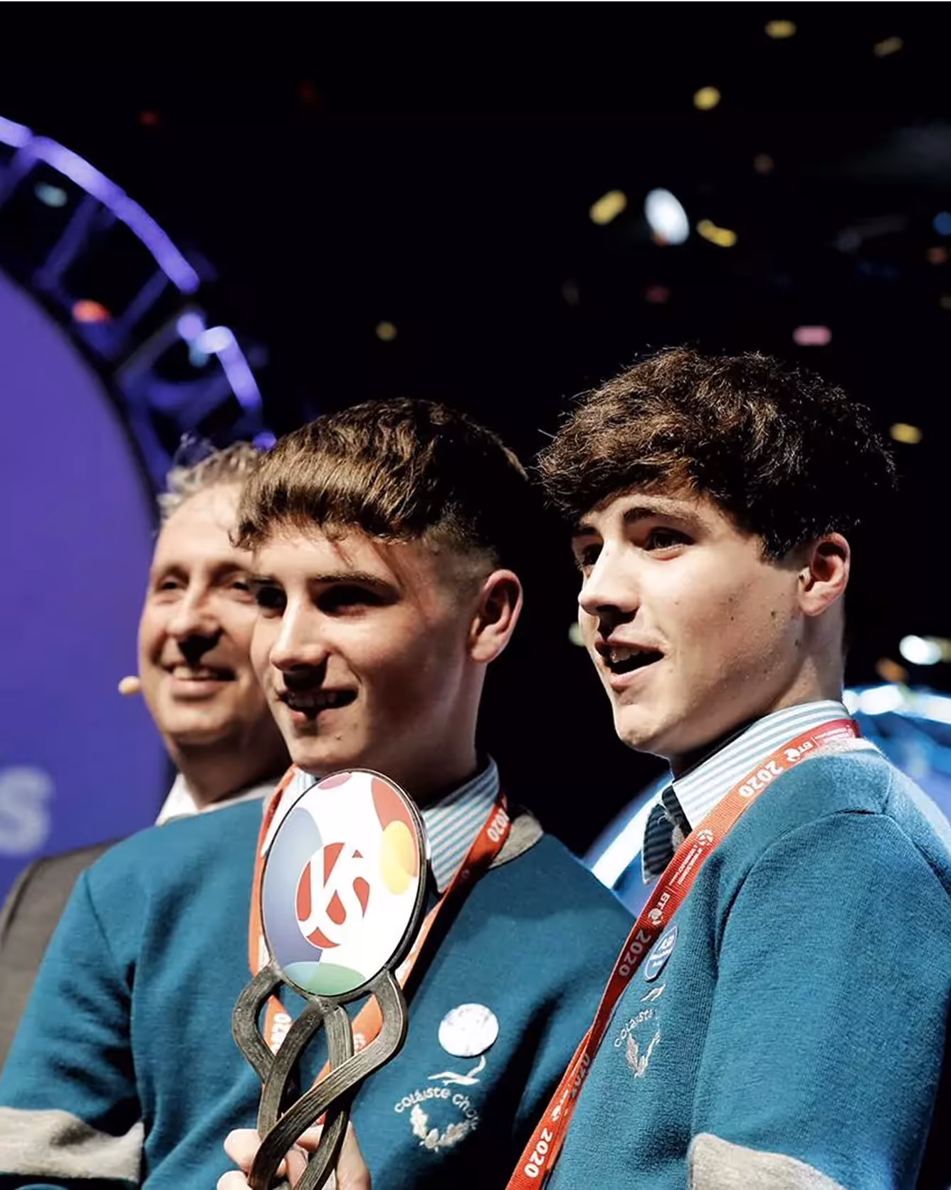 Two students in blue uniforms accepting award trophy on stage.