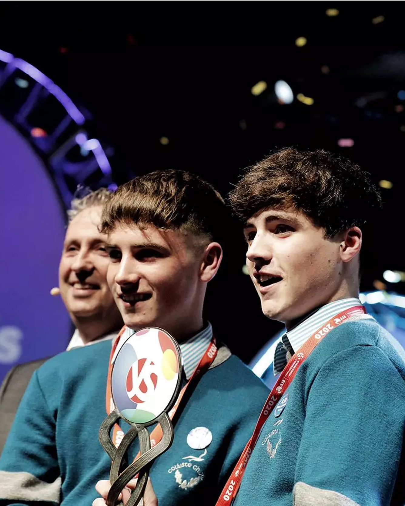 Two students in blue uniforms accepting award trophy on stage.