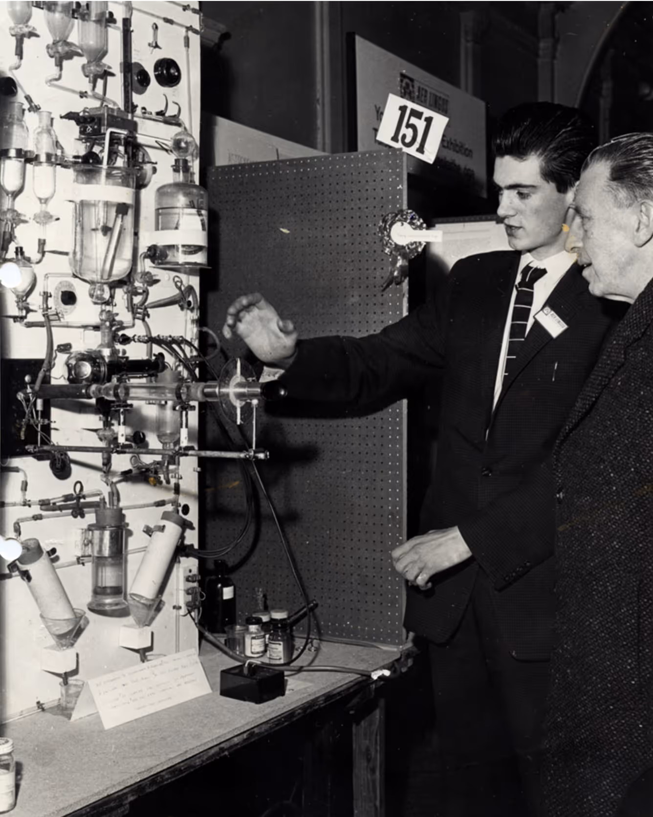 Young man in suit demonstrating a science experiment setup to an older man at an exhibition.