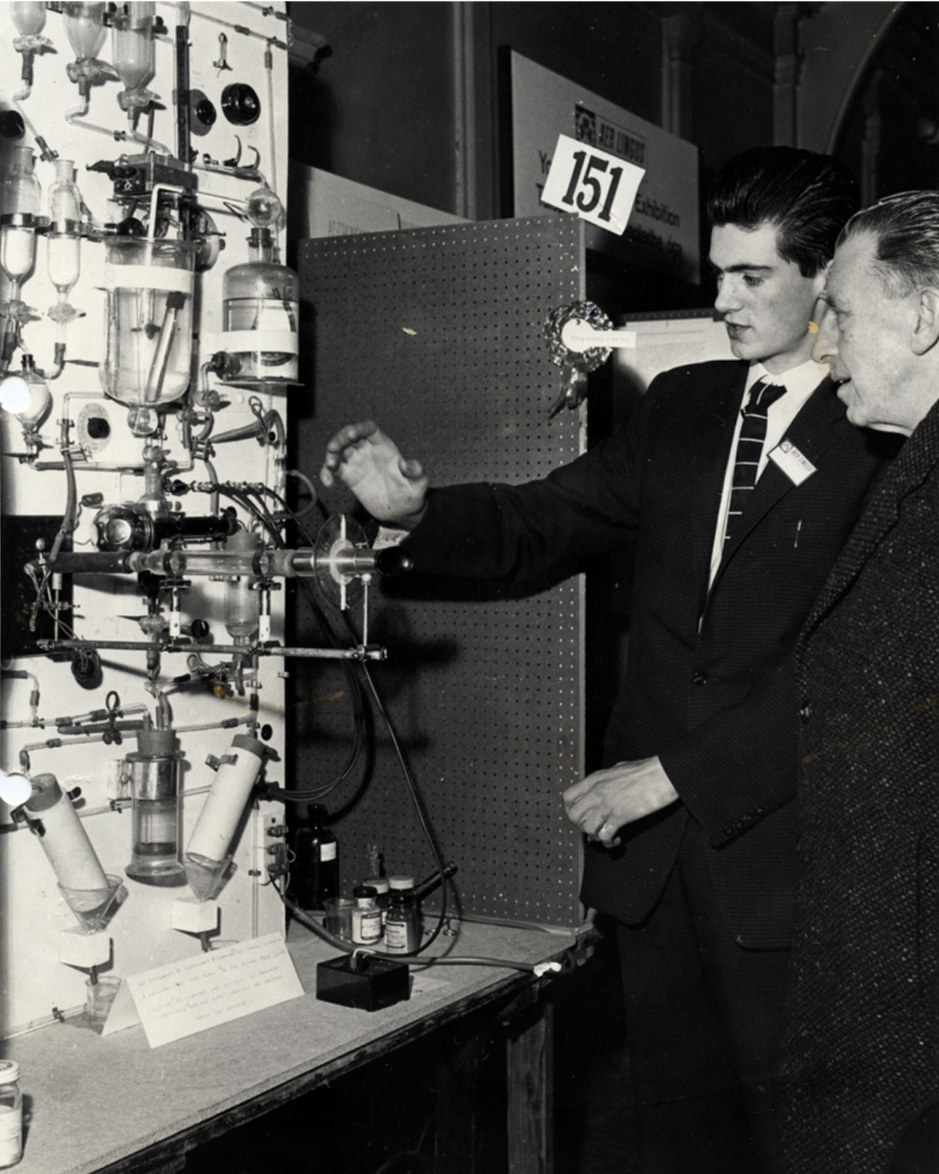 Young man in suit demonstrating a science experiment setup to an older man at an exhibition.