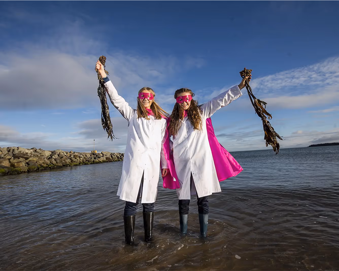 Two girls in lab coats and pink capes standing in water holding seaweed.
