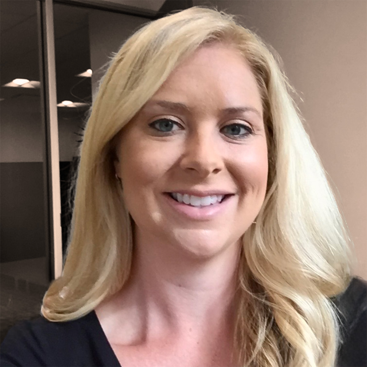 A smiling woman with long blonde hair poses for a portrait in a modern office setting.